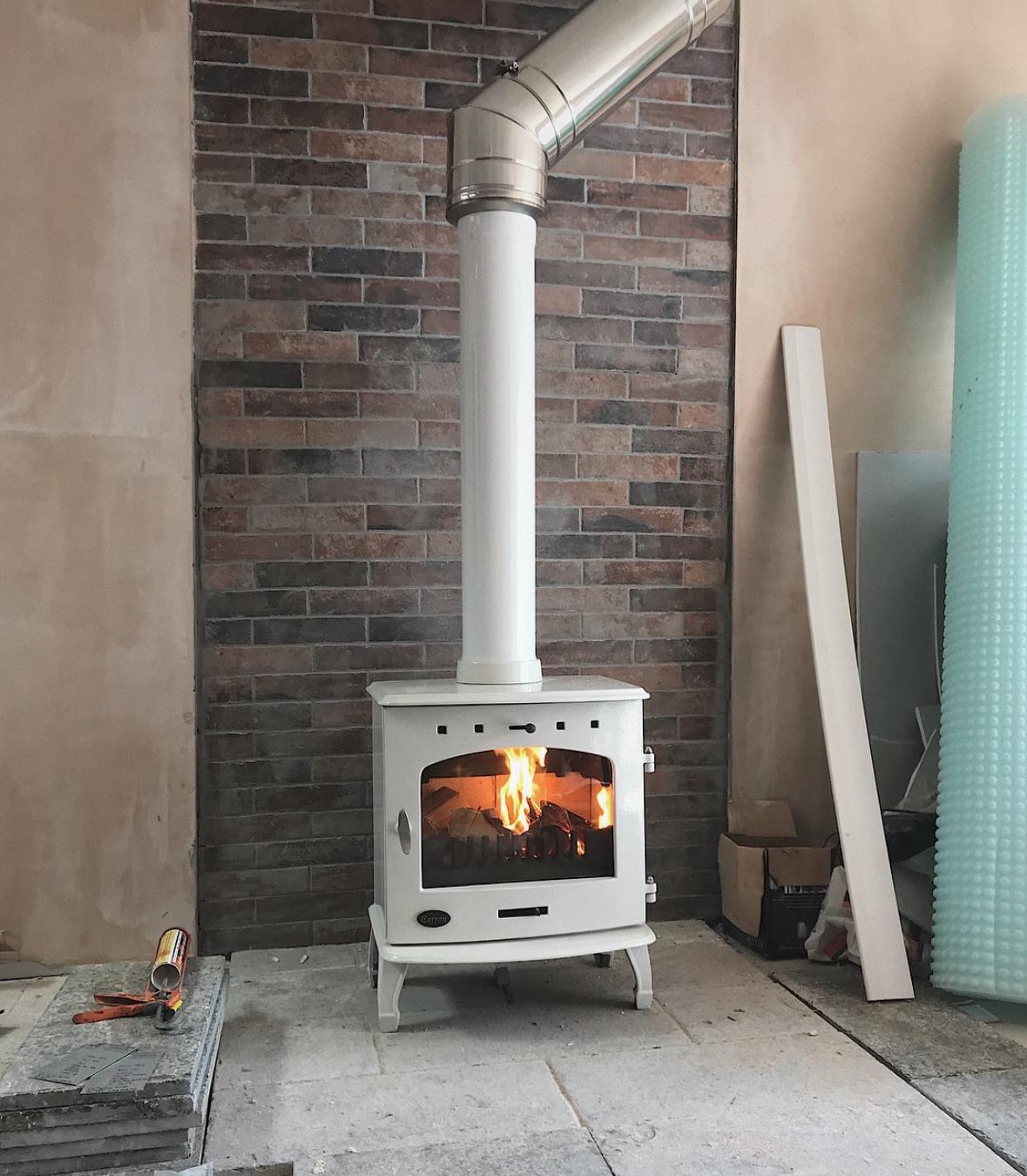 A small white wood-burning stove with a visible fire inside, connected to a chimney pipe, situated against a brick wall.