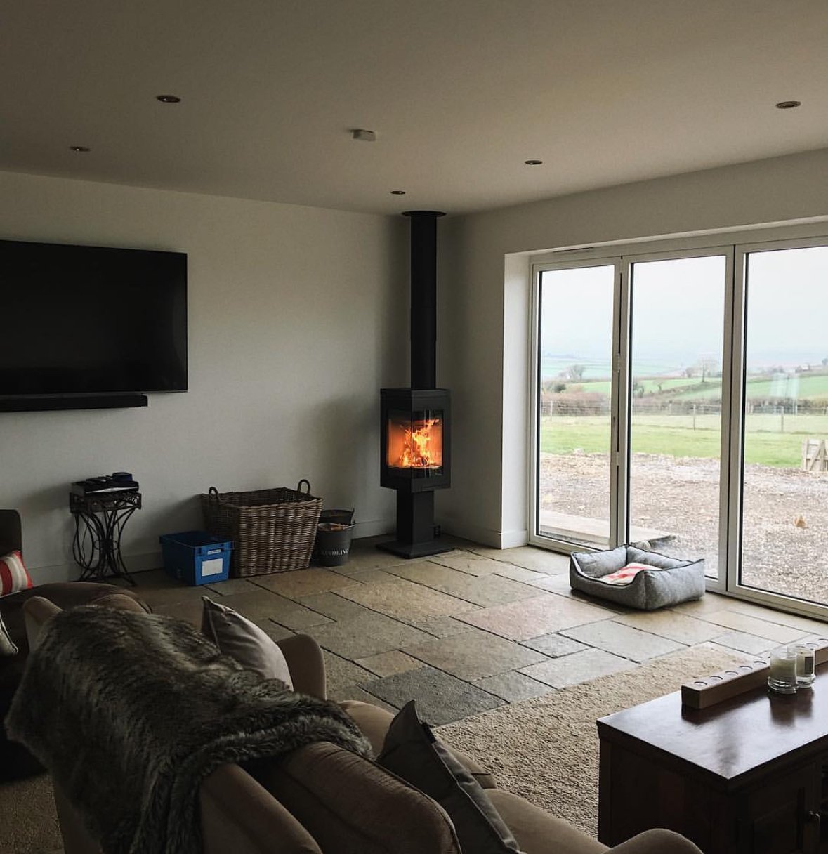 Living room with a wall-mounted TV, a wood stove with a fire, sliding glass doors leading outside to a rural landscape, and a pet bed on the floor.