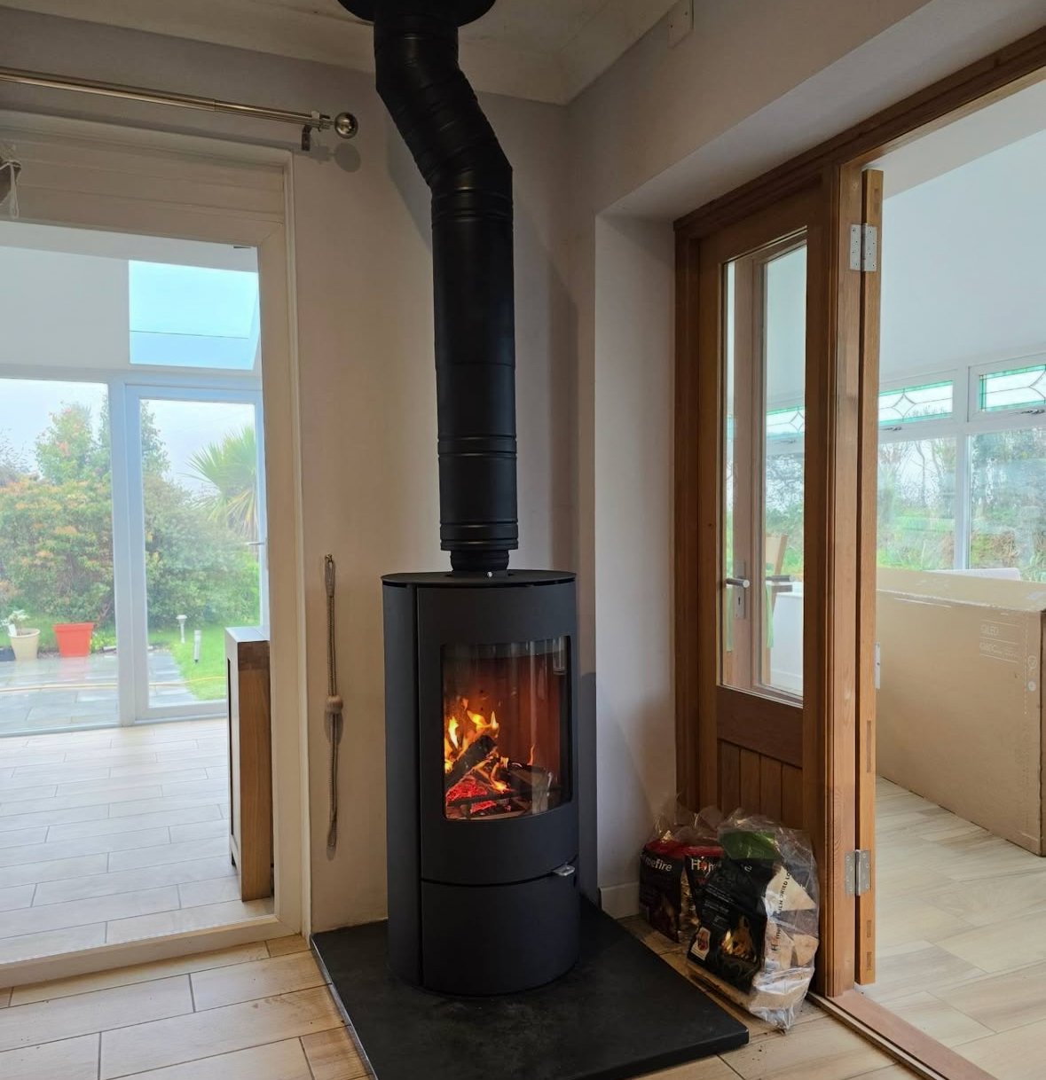 Black wood stove with a glass door in a living room, burning logs inside, with a black pipe extending upward through the ceiling. To the right, a glass door leads to a porch, and a bag of firewood is on the floor beside it.