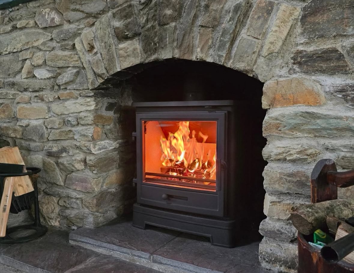 A wood stove with a glass door showing a bright fire inside, set in a stone fireplace. The fireplace is constructed of irregularly shaped stones and has a curved arch at the top.