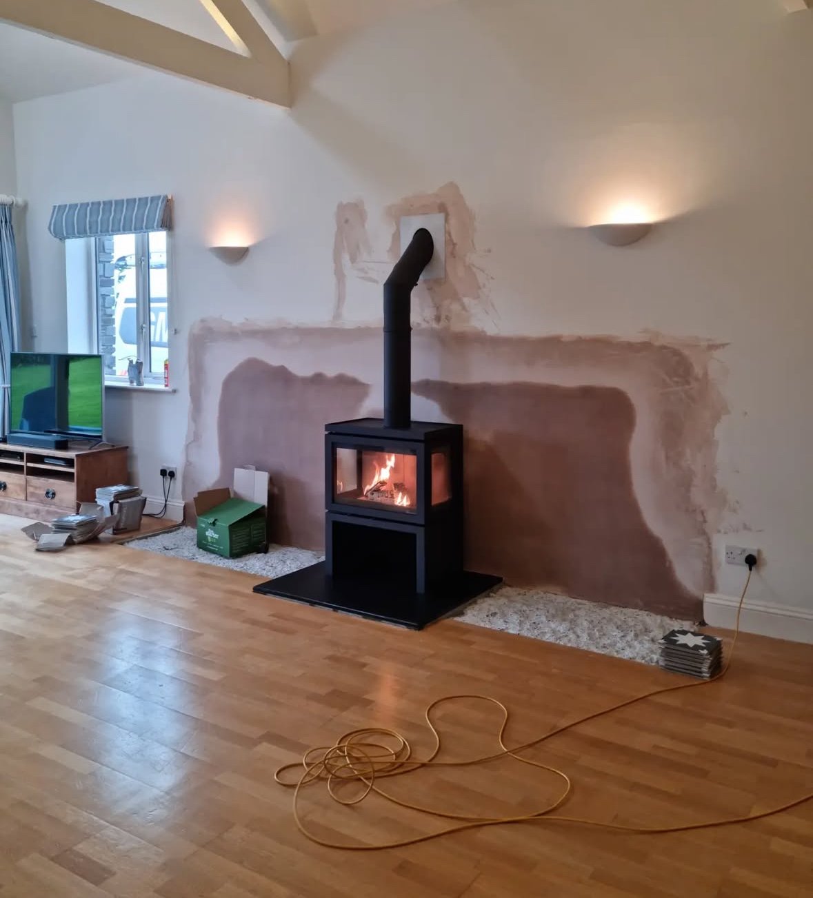 Living room with a stovepipe fireplace surrounded by unfinished wall patching, a window with striped curtains, a television on a wooden stand, and various items on the floor, including an extension cord and a green cardboard box.