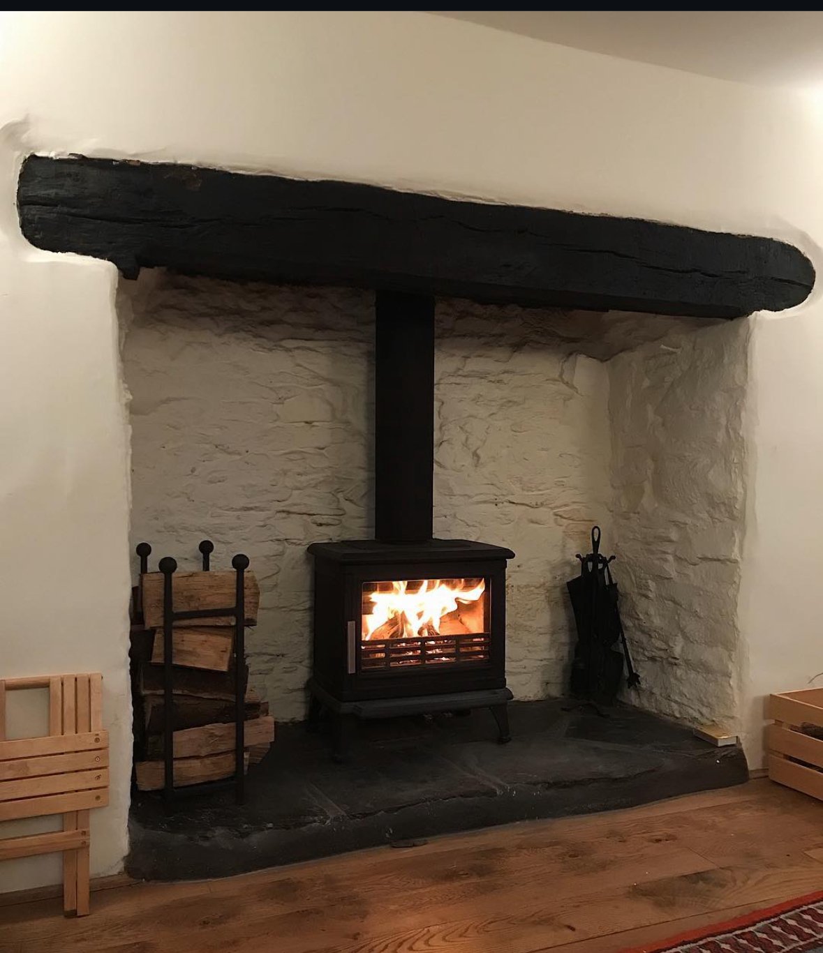 A cozy room featuring a wood-burning stove with flames, set against white stone walls and a black wooden mantel. Firewood is stored in a rack on the left and fire tools are on the right.