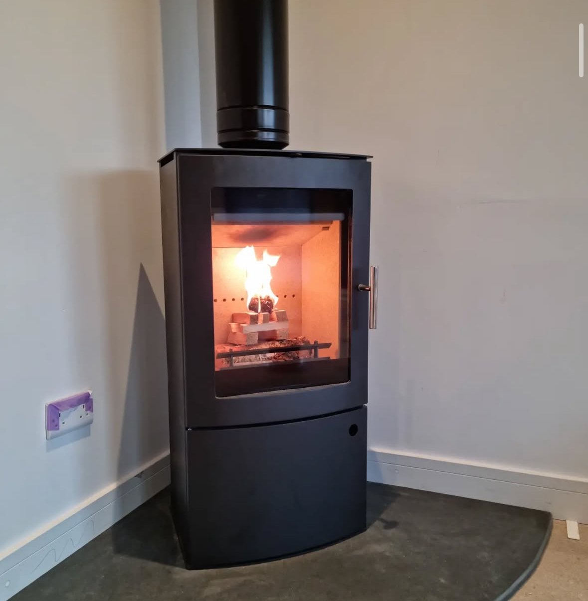 Black wood stove with glass door showing a fire inside, located against a plain white wall in a room with gray flooring.