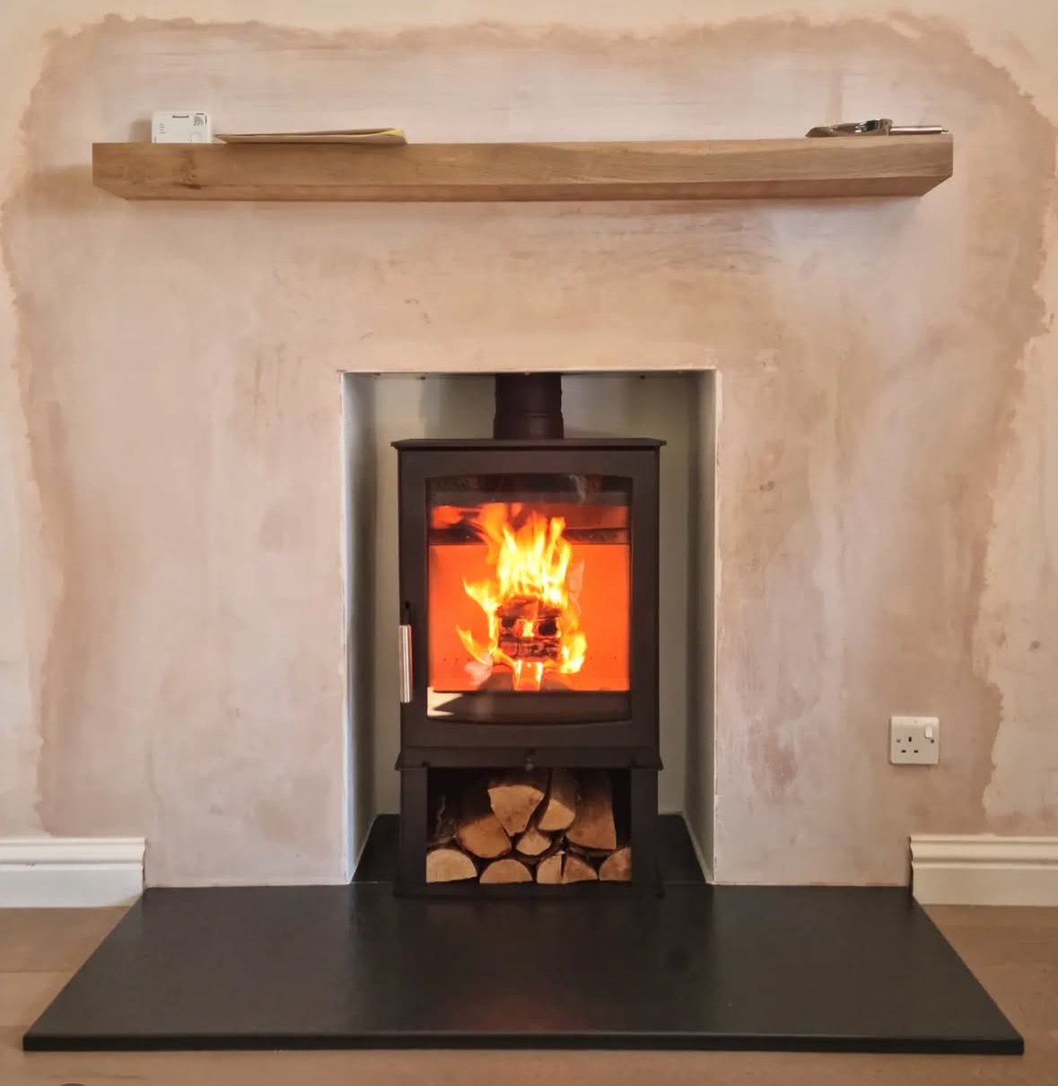 A wood-burning stove with fire inside, set in a recessed wall with a wood fire storage beneath, and a wooden mantle shelf above, in a living room.