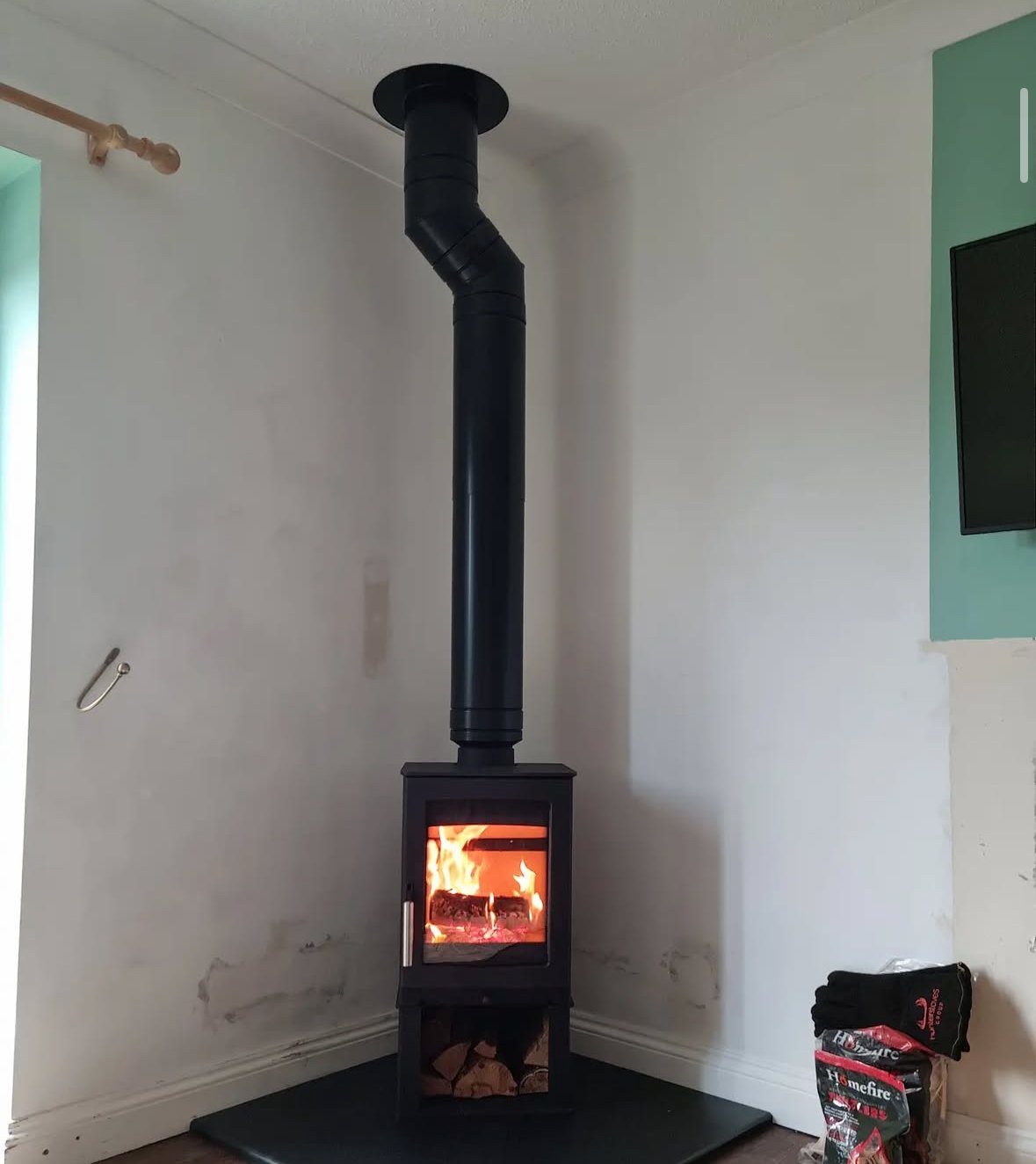 A black wood stove with a glass door showing a fire inside, attached to a black chimney pipe extending to the ceiling, situated in a corner of a room with white walls and a pile of firewood underneath.