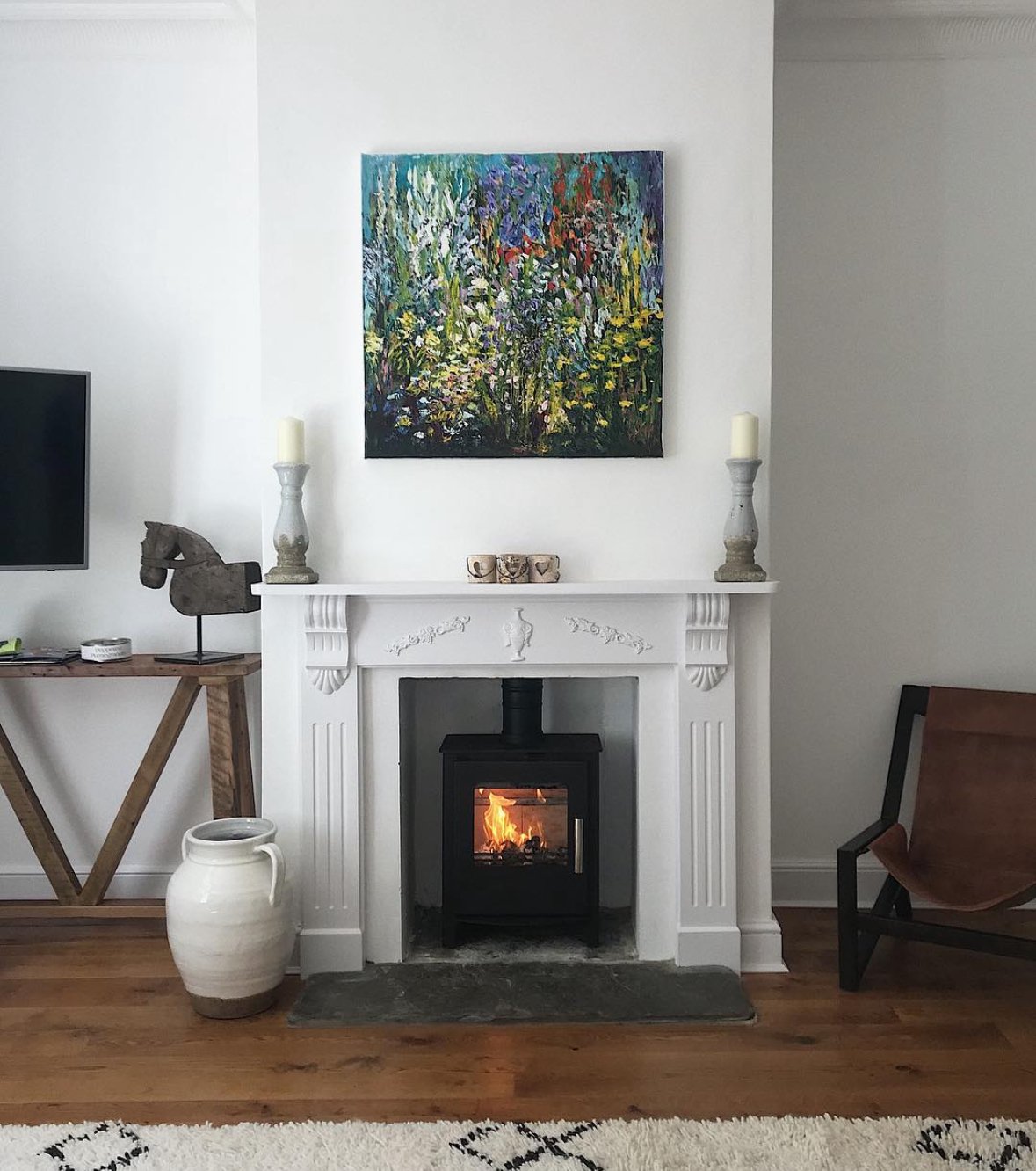 Living room with a white fireplace, a colorful abstract painting above, a wood stove with fire burning, with decorative candles and small ceramics on the mantel, a large white vase on the floor, a wooden chair with leather cushion on the right, and p