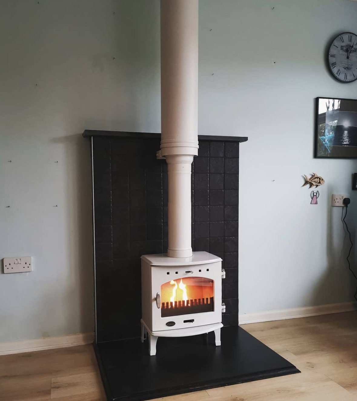 White wood-burning stove with a glowing fire, set against a black tile backdrop on a black hearth in a living room.