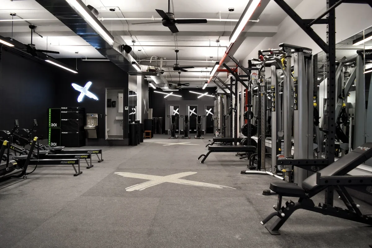 Empty modern gym with strength and cardio equipment, black walls, and a neon X sign.