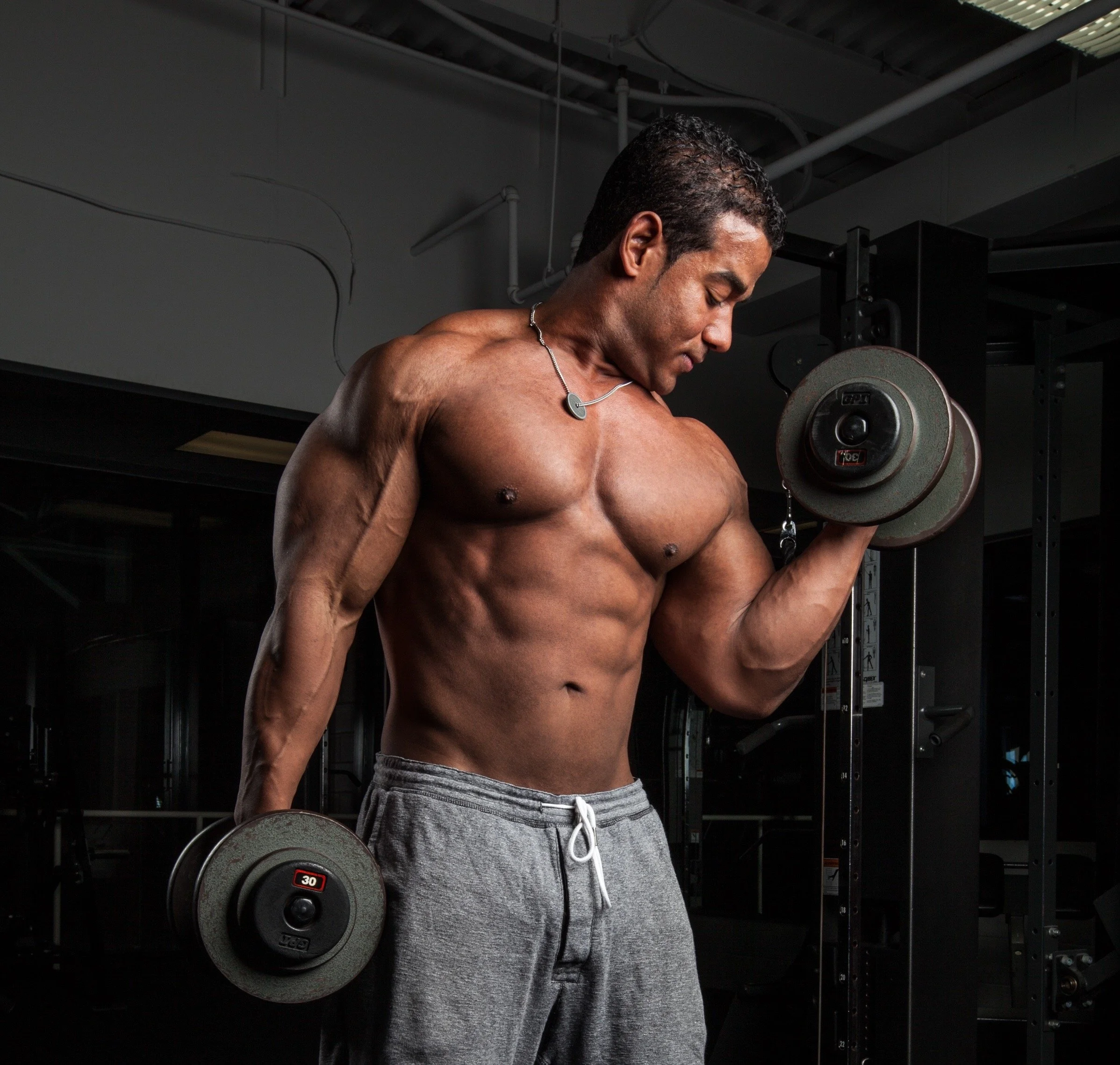 A muscular man lifting dumbbells in a gym, wearing gray sweatpants and a necklace, with a focused expression.