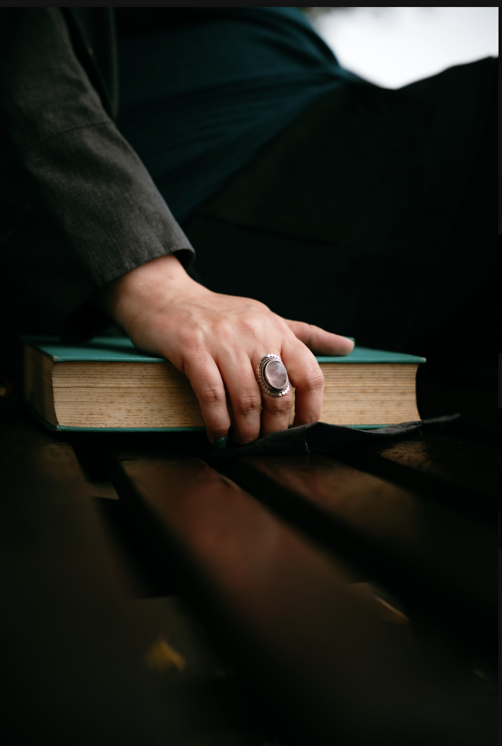 A hand resting on a closed book on a dark wooden surface, wearing a large silver ring with a gemstone.