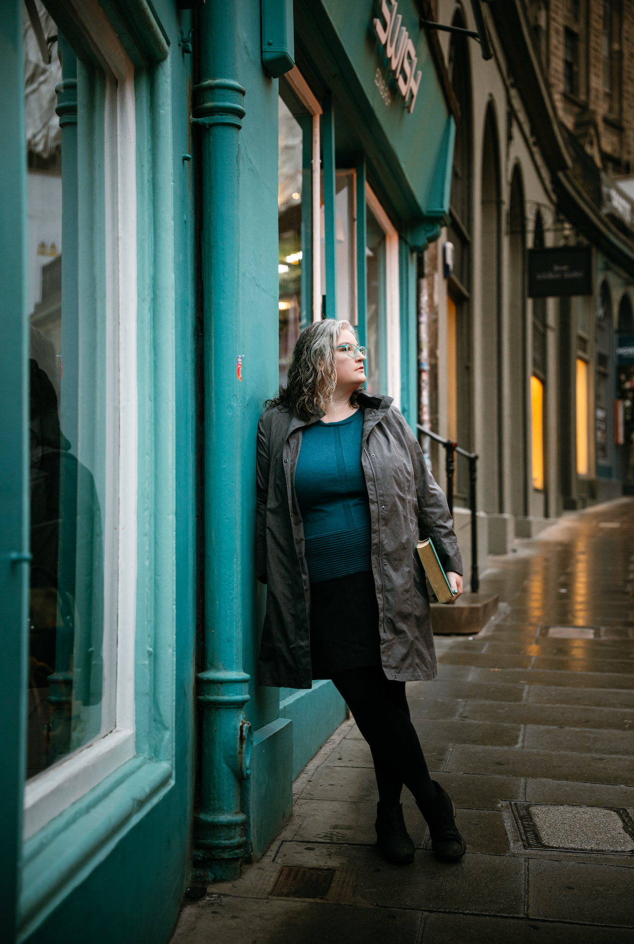 A woman with curly gray hair, glasses, wearing a dark coat and black pants, stands against a teal storefront on a city street, holding a book or folder, with buildings and shops in the background.