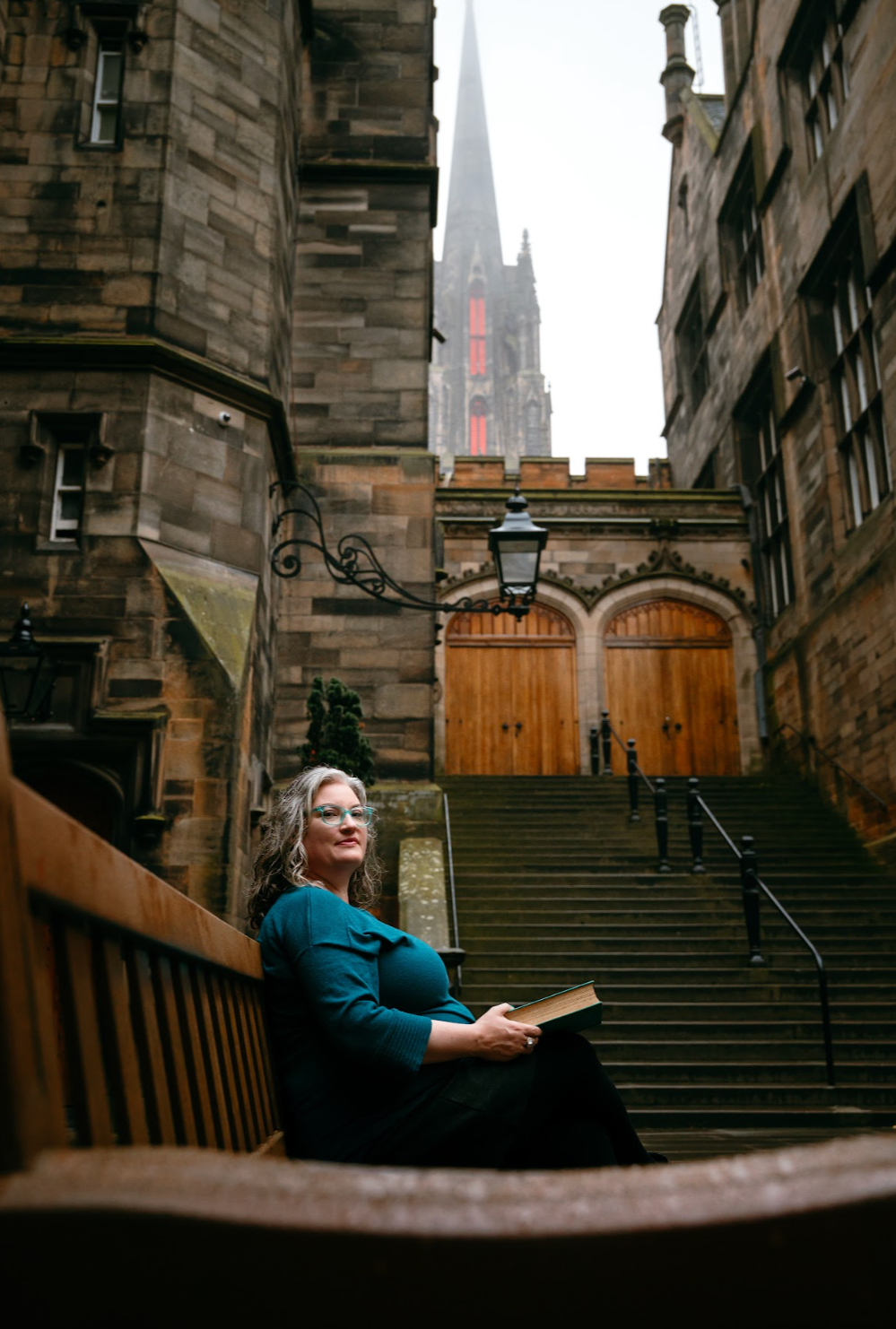 A woman with glasses and curly gray hair sitting on a wooden bench outside, holding a book, in front of stone stairs leading up to wooden doors, with an old stone building and a tall church spire in the background.