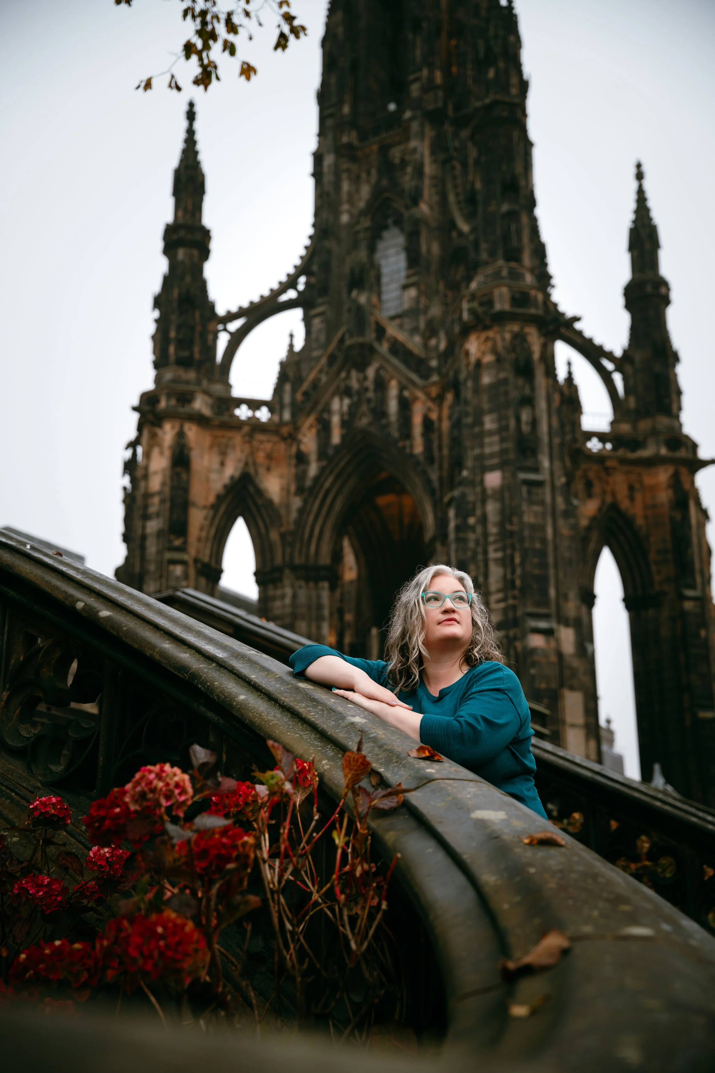 A woman with glasses and curly gray hair leaning on a black wrought iron railing, with a Gothic cathedral tower in the background on an overcast day.