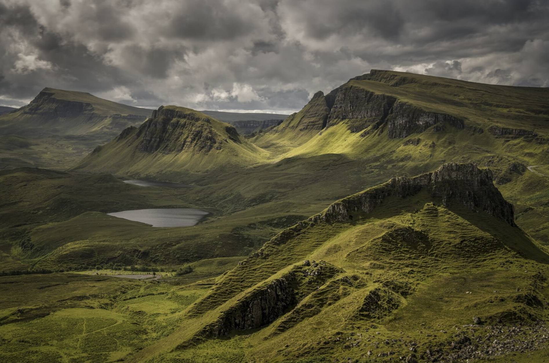 A scenic landscape featuring green rolling hills and steep mountain cliffs, with dark, brooding clouds overhead and a reflective lake in the valley.