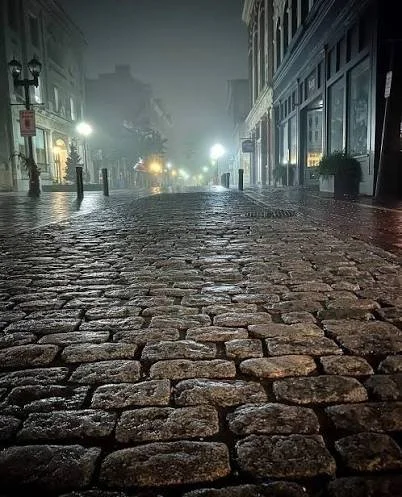 Empty cobblestone street at night with buildings, streetlights, and foggy atmosphere.
