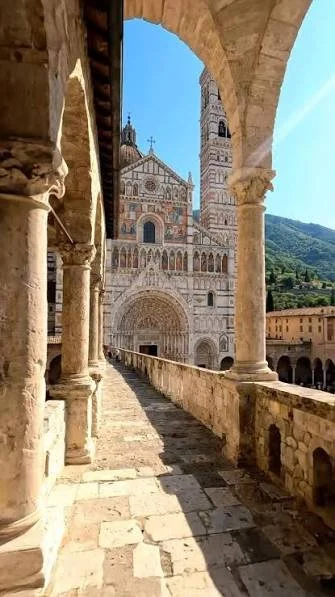 View through archways of historic stone cathedral with detailed facade, bell tower, and surrounding lush green hills under bright blue sky.