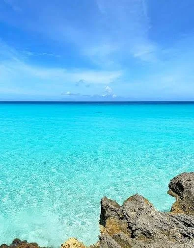 Turquoise ocean water with rocks in the foreground, clear blue sky with a few clouds in the background.