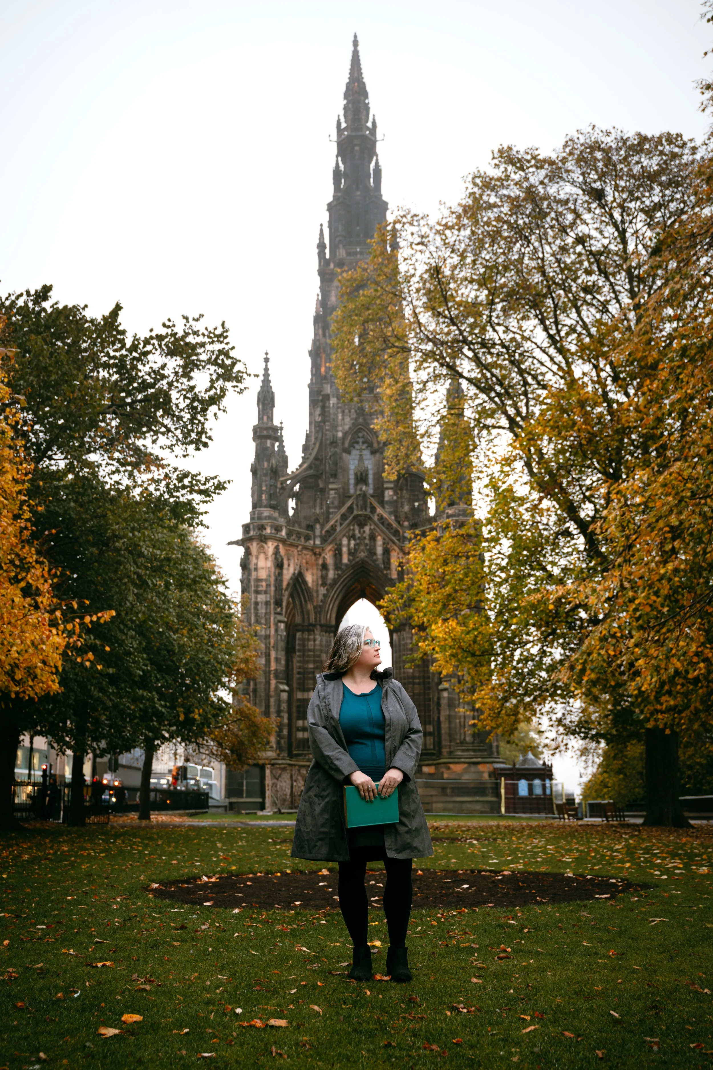 A woman standing on a grassy area with fallen autumn leaves, wearing a grey coat and holding a turquoise folder, in front of an ornate gothic-style church tower, surrounded by trees with autumn foliage.