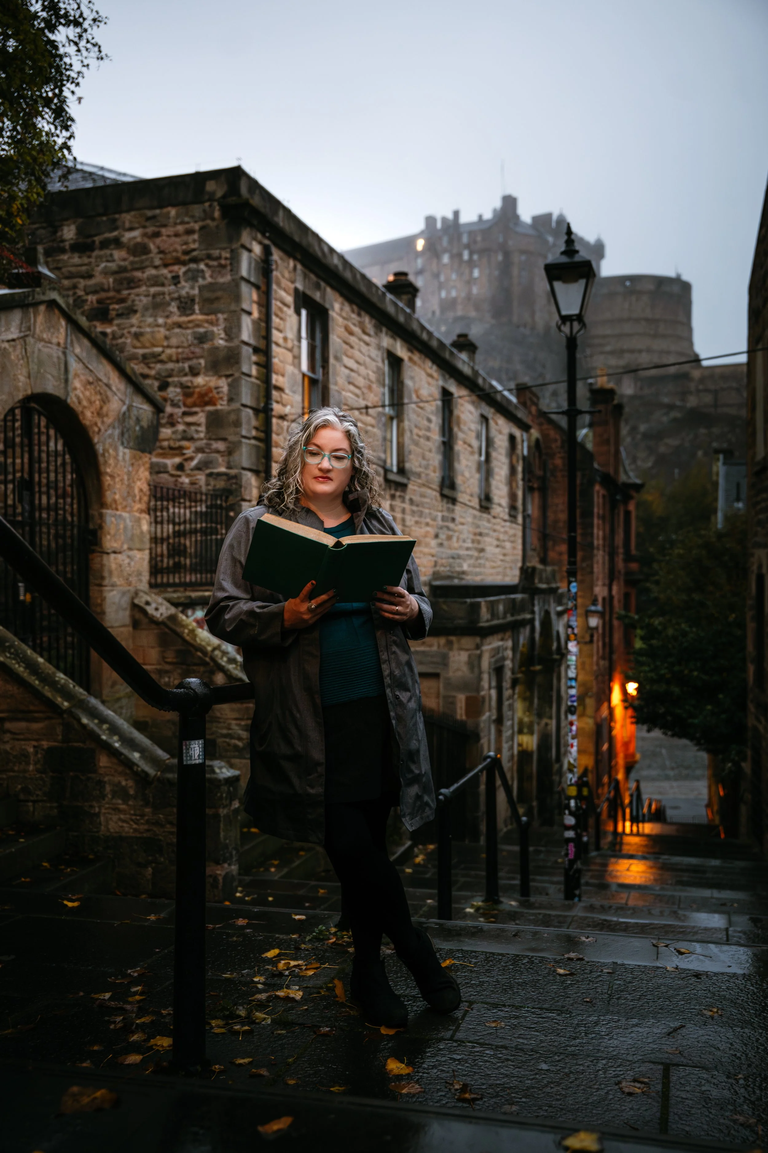 A woman with curly gray hair, glasses, and a dark coat stands on wet stairs holding a green book, with an old stone building and a castle in the background during dusk or early evening, with autumn leaves on the ground.