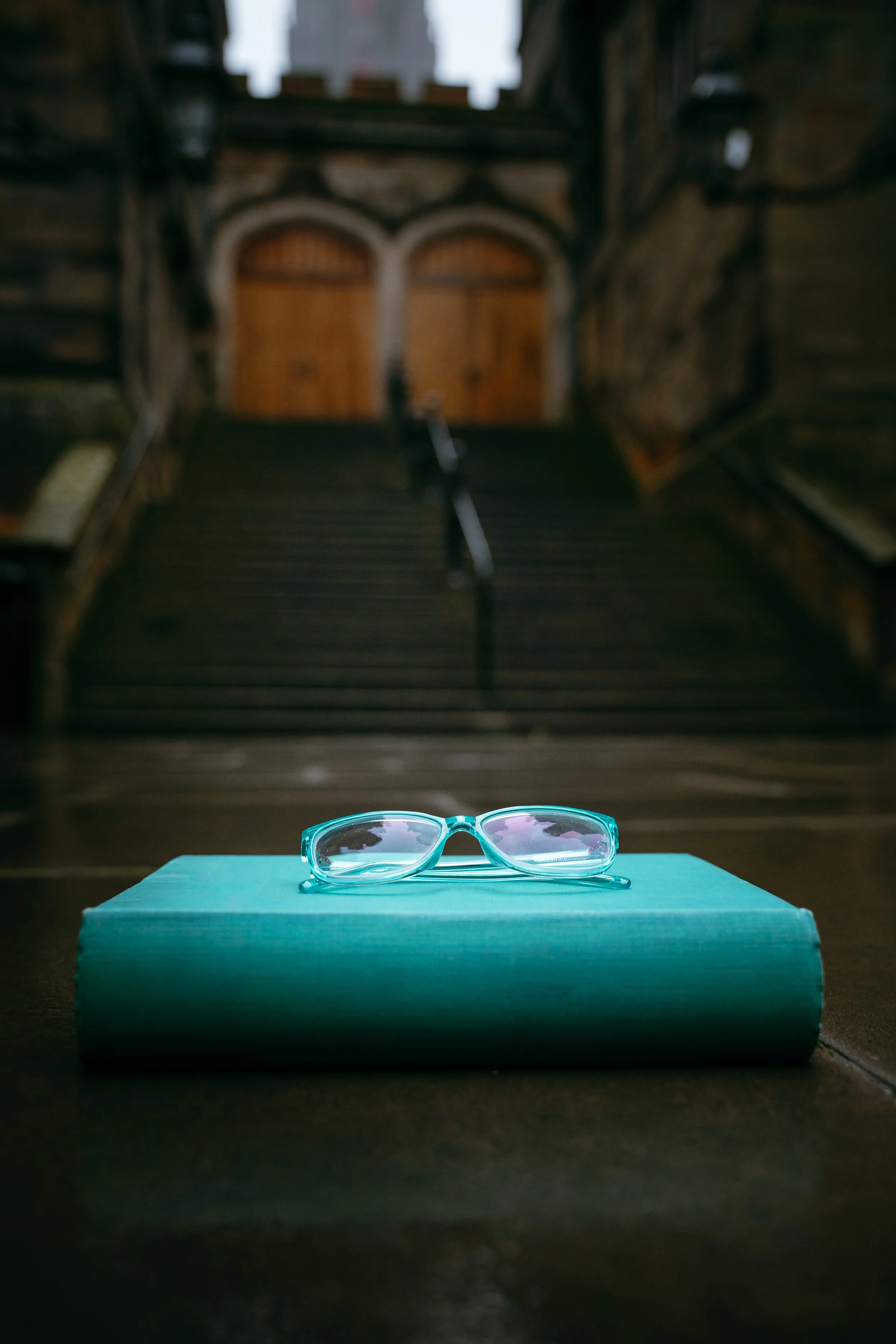 A pair of turquoise eyeglasses resting on a closed teal book, with a background of a wooden staircase and doors.