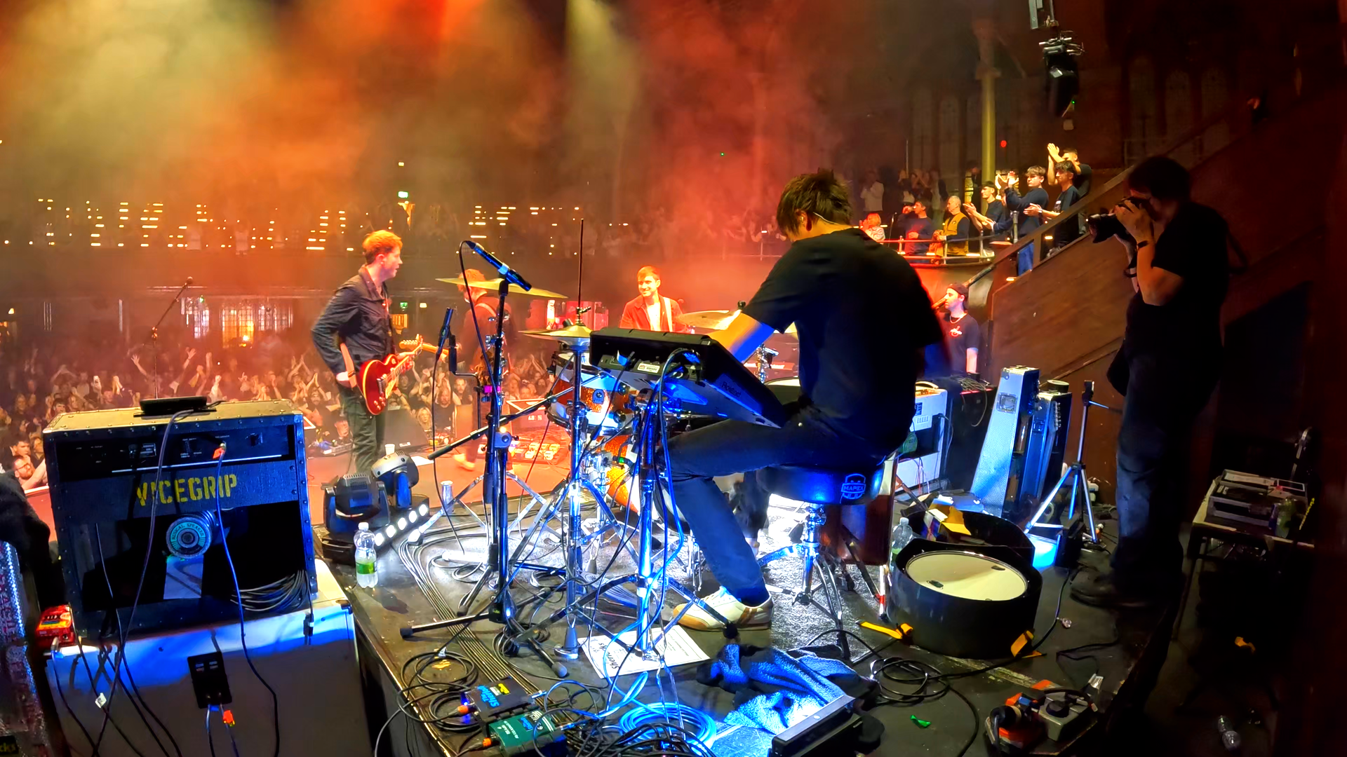 Band performing live on stage at a concert, with a crowd of fans in front, colorful stage lighting, and musicians playing guitars and keyboards.