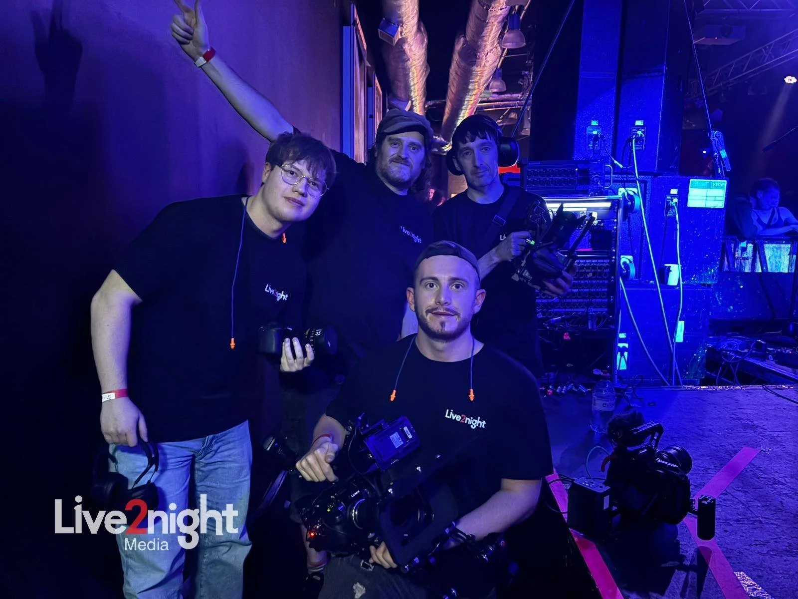 Four media crew members at a concert or event backstage area with equipment, blue lighting, and a stage in the background, wearing black 'Live2night' branded shirts, some holding cameras and headphones.