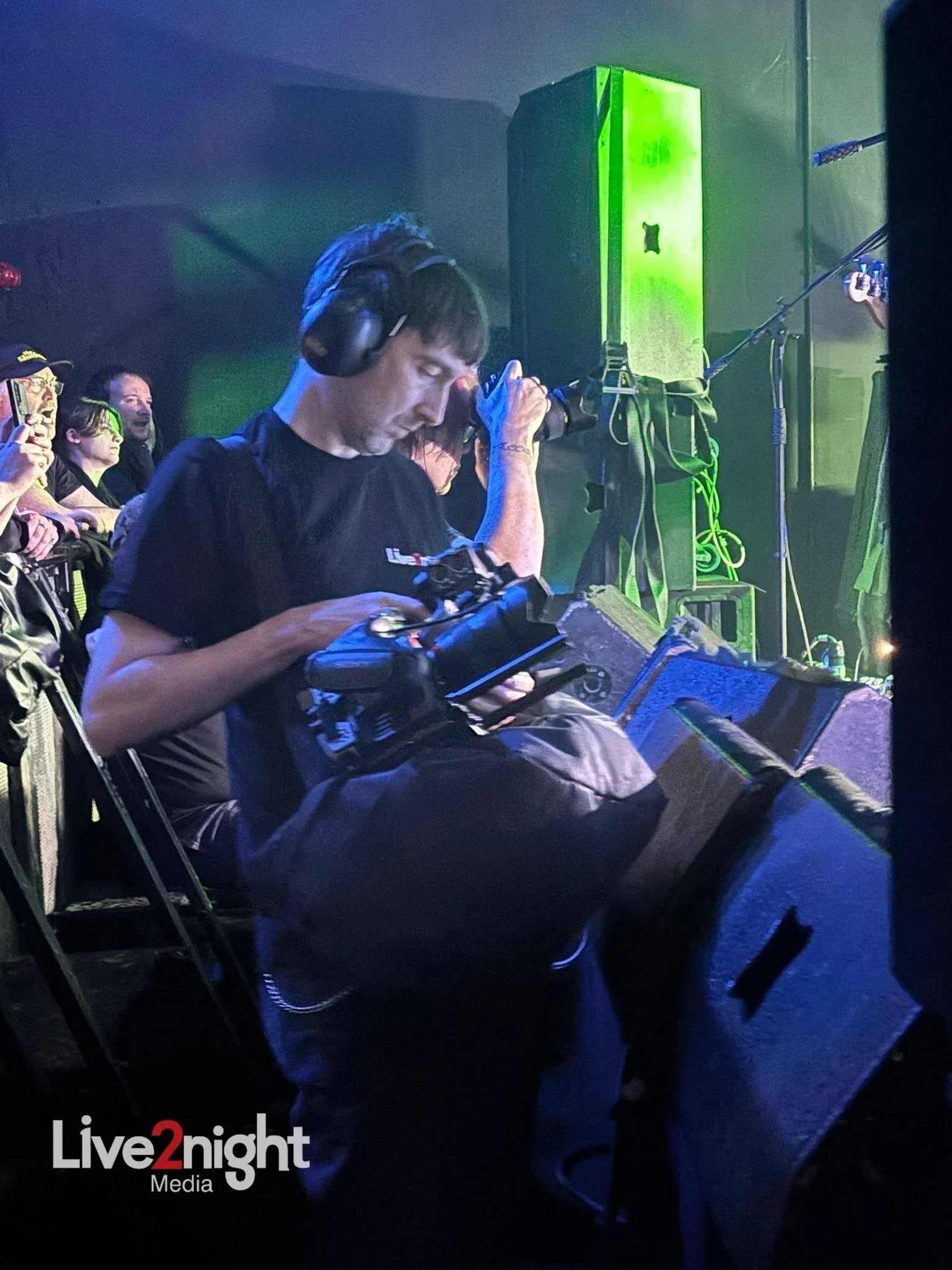 A man wearing headphones, holding a camera, and looking down at it, sitting near a large speaker at a live music event with a crowd in the background and bright green stage lighting.