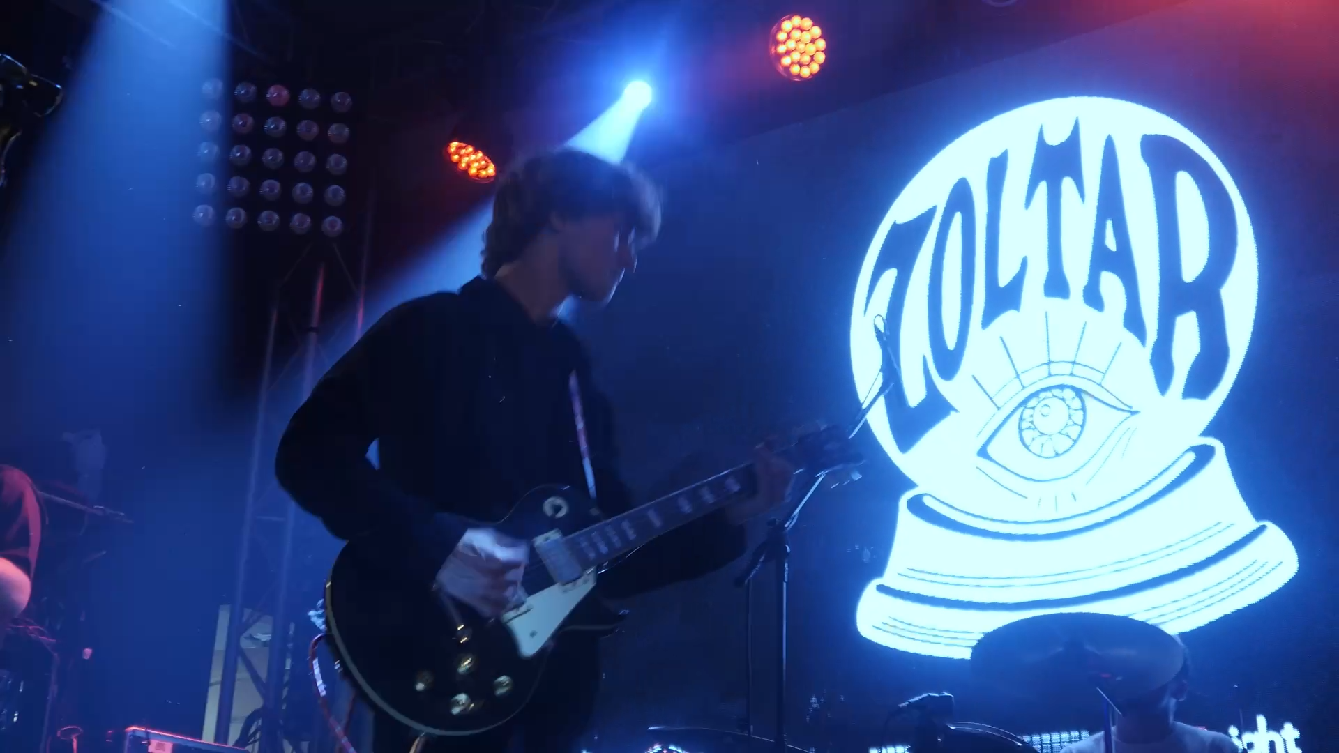 A musician playing an electric guitar on stage with colorful lights and a large illuminated logo that says 'LOSTHOUR' with an eye inside a globe symbol.