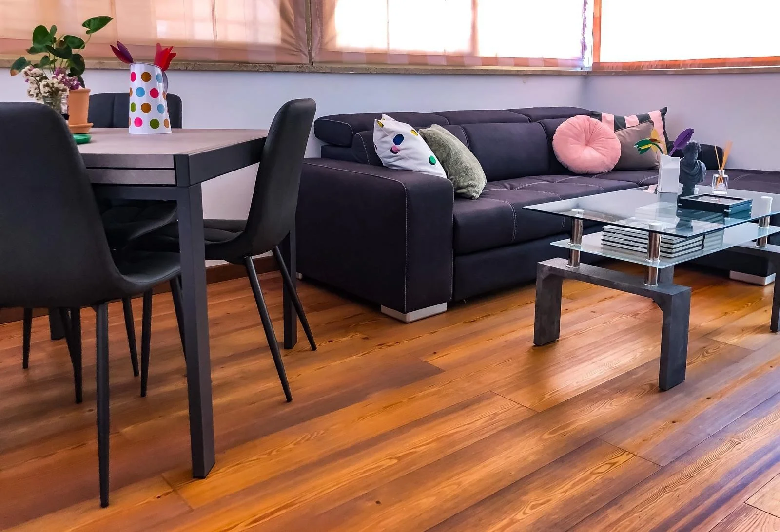 Soggiorno con divano nero, tavolino in vetro e sedie nere, decorato con cuscini colorati e piante, pavimento in legno.