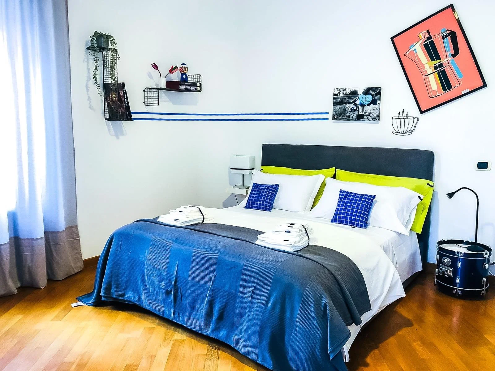 Camera da letto moderna con letto matrimoniale, lenzuola bianche, cuscini blu a quadretti e una coperta grigia. Sul letto ci sono due asciugamani ripiegati. A destra, lampada da tavolo nera e su una piccola mensola di fianco un orologio da parete e u