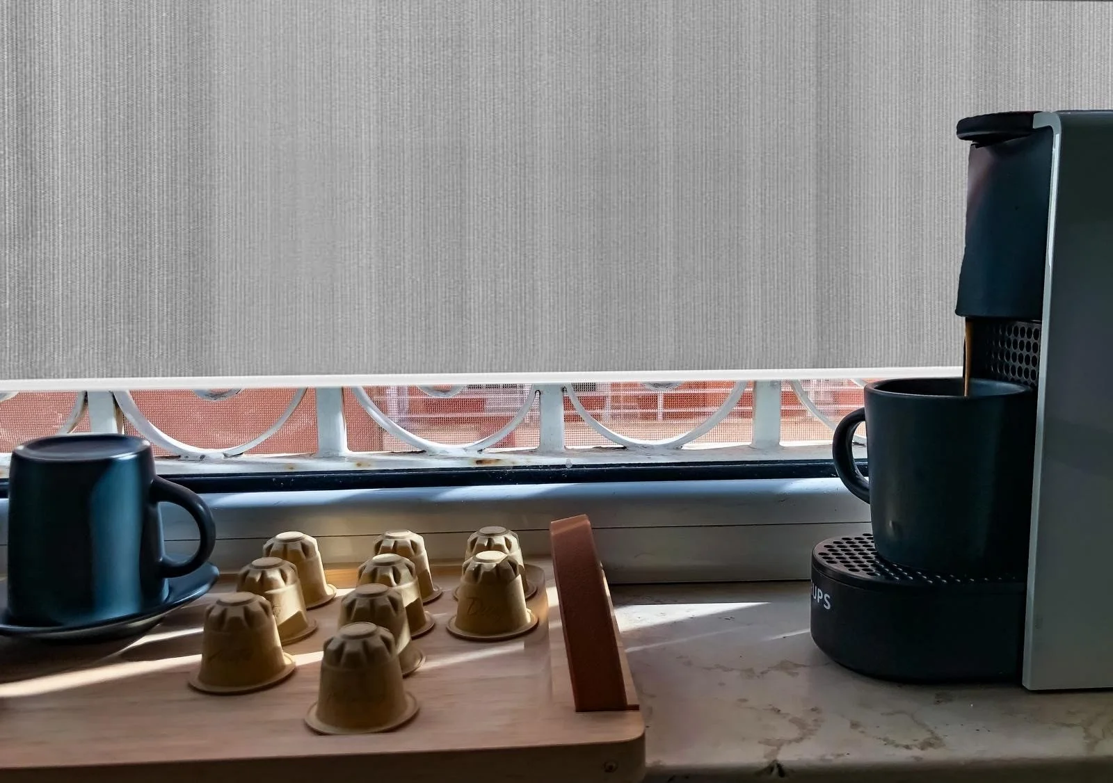Macchina da caffè con tazza, cialde di caffè e tazza di caffè sul davanzale di una finestra con una copertura a metà e ringhiera.