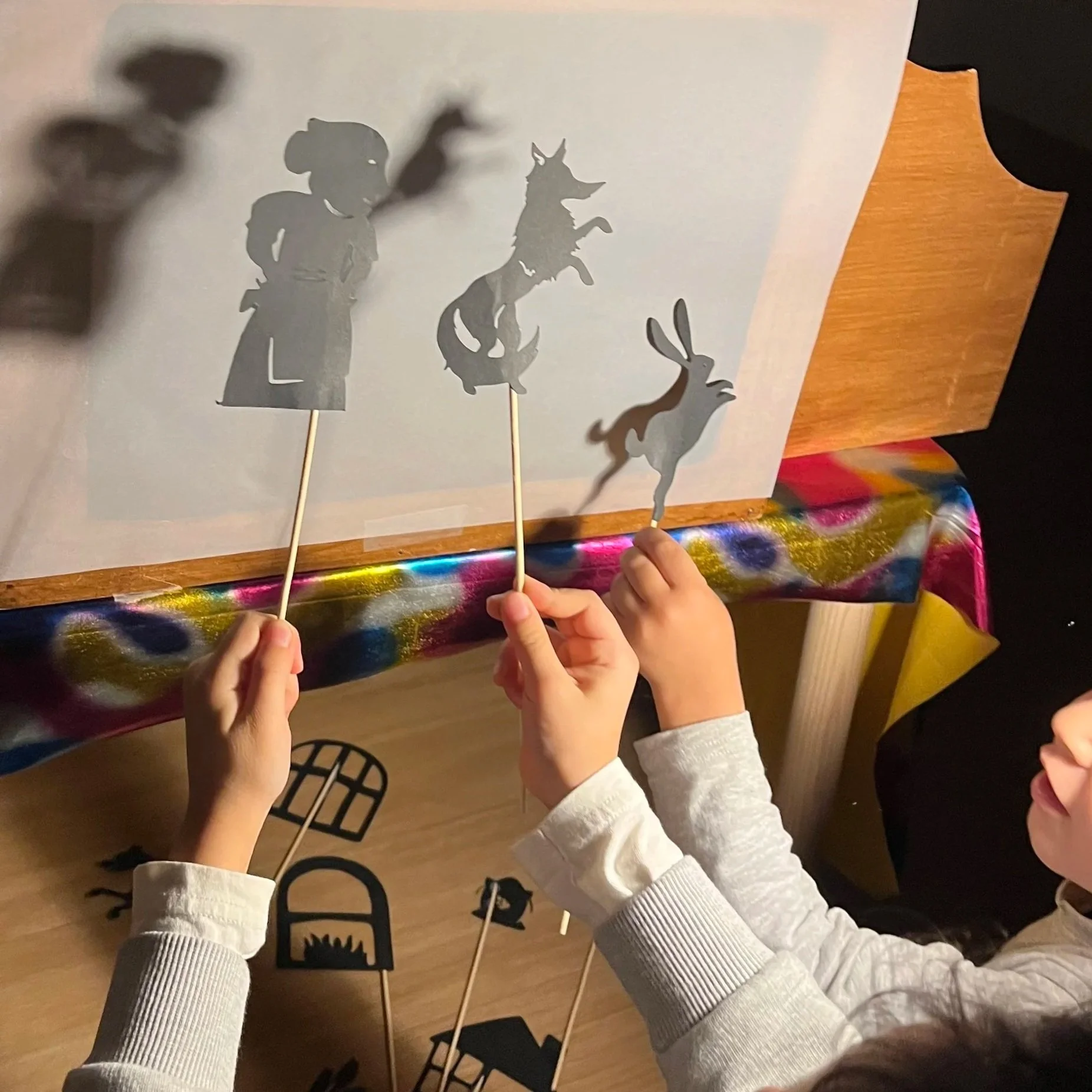 Une personne tient un bâton avec une silhouette de lapin en carton, créant une ombre en projection sur un mur blanc. D'autres silhouettes en carton, comme un personnage avec un chapeau, un dragon, et une maison, sont également alignées sur une table 