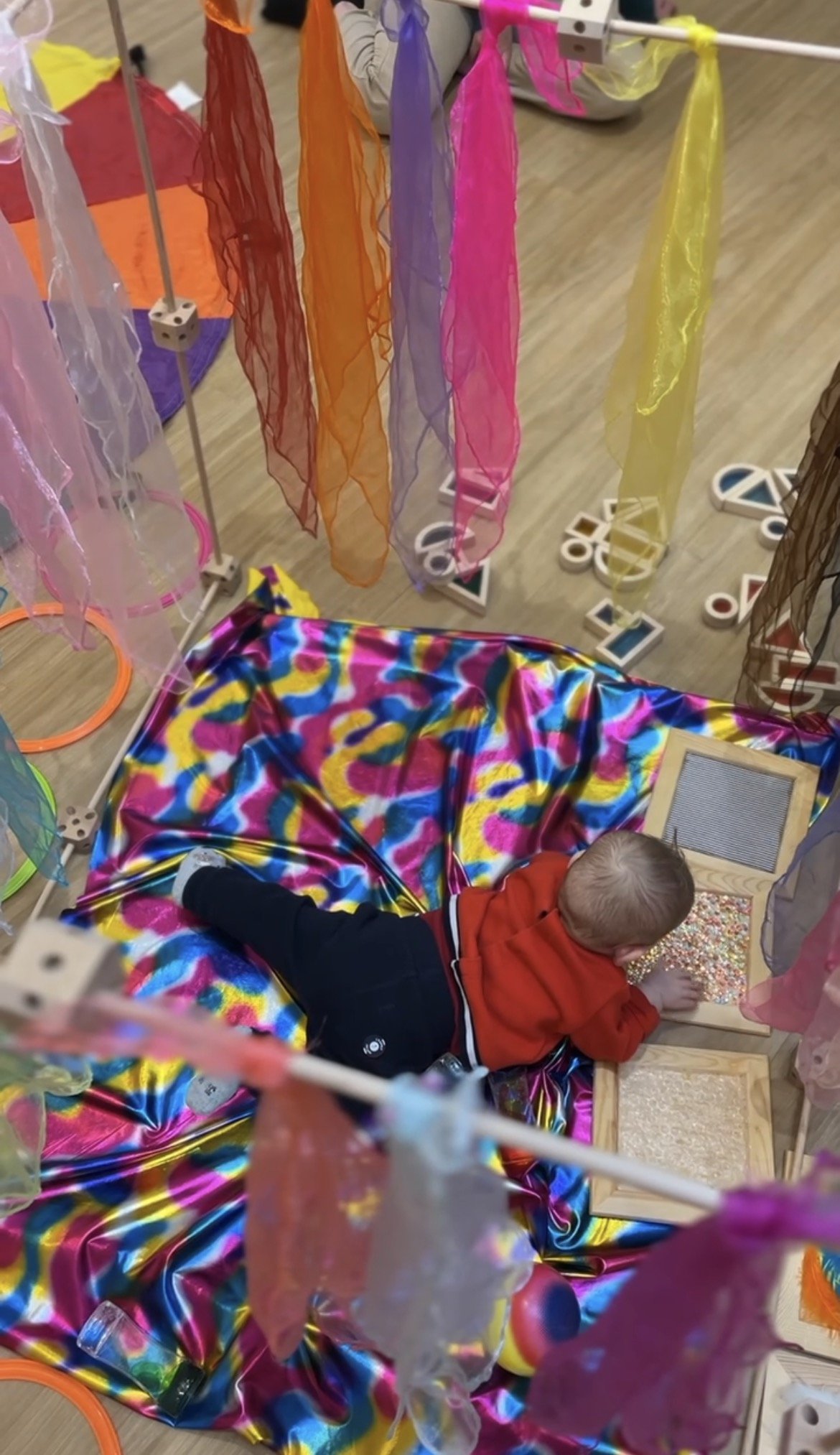 Un jeune enfant allongé sur une couverture colorée, jouant avec des perles dans un espace de jeu coloré.