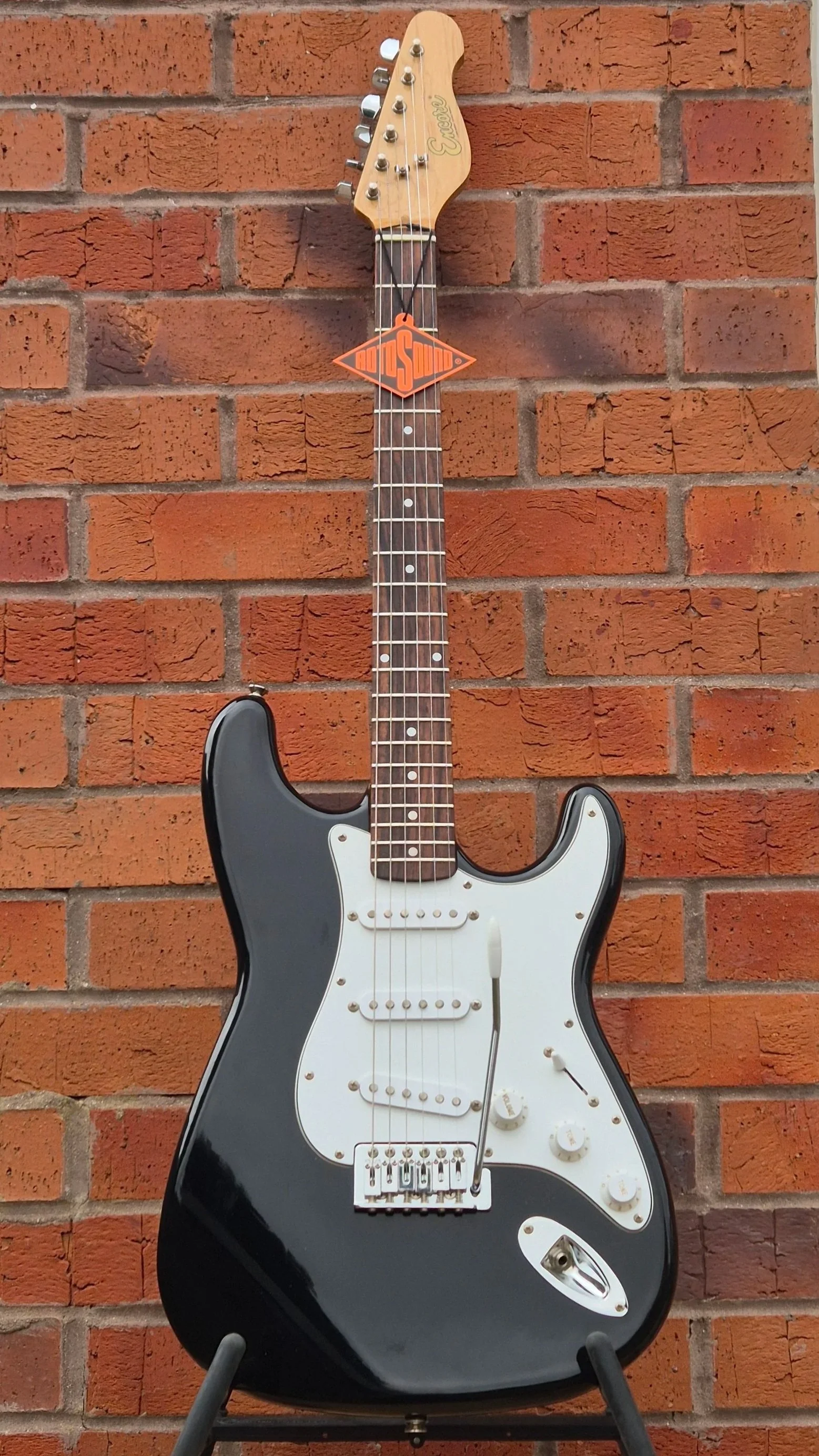 A black electric guitar with a white pickguard, mounted on a stand against a red brick wall.