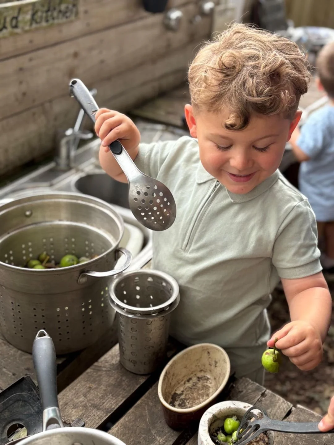 A young boy with curly hair in a light-colored shirt is smiling and holding a small green fruit, possibly an olive, with a fork. He is outdoors, near a wooden fence, with a metal strainer, various utensils, and small bowls of green fruits on a rustic wooden table.