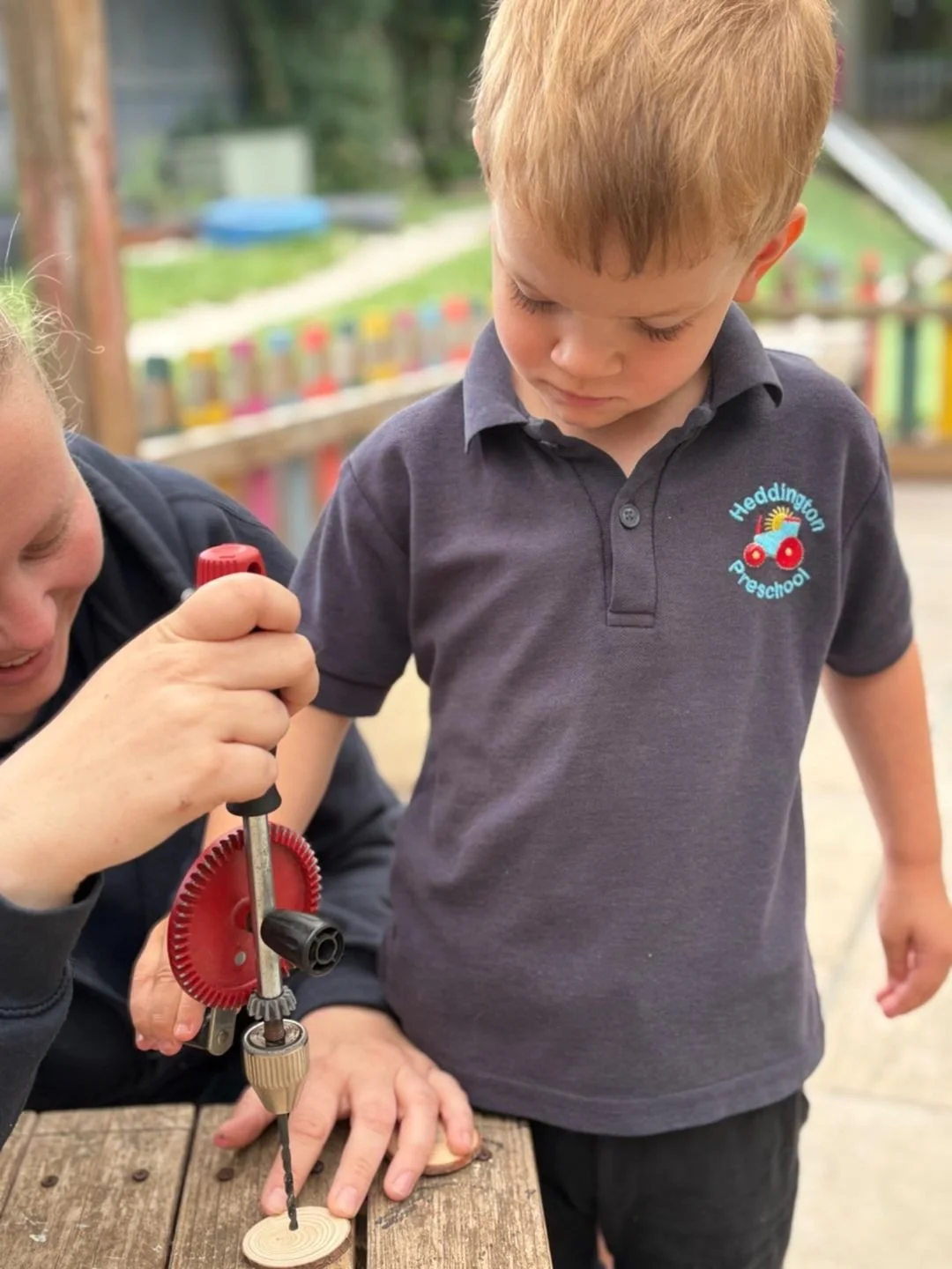 A young boy with red hair wearing a navy blue polo shirt with a preschool logo is watching a woman operate a drill on a small wooden piece outdoors. The background shows a colorful, blurred fence and greenery.