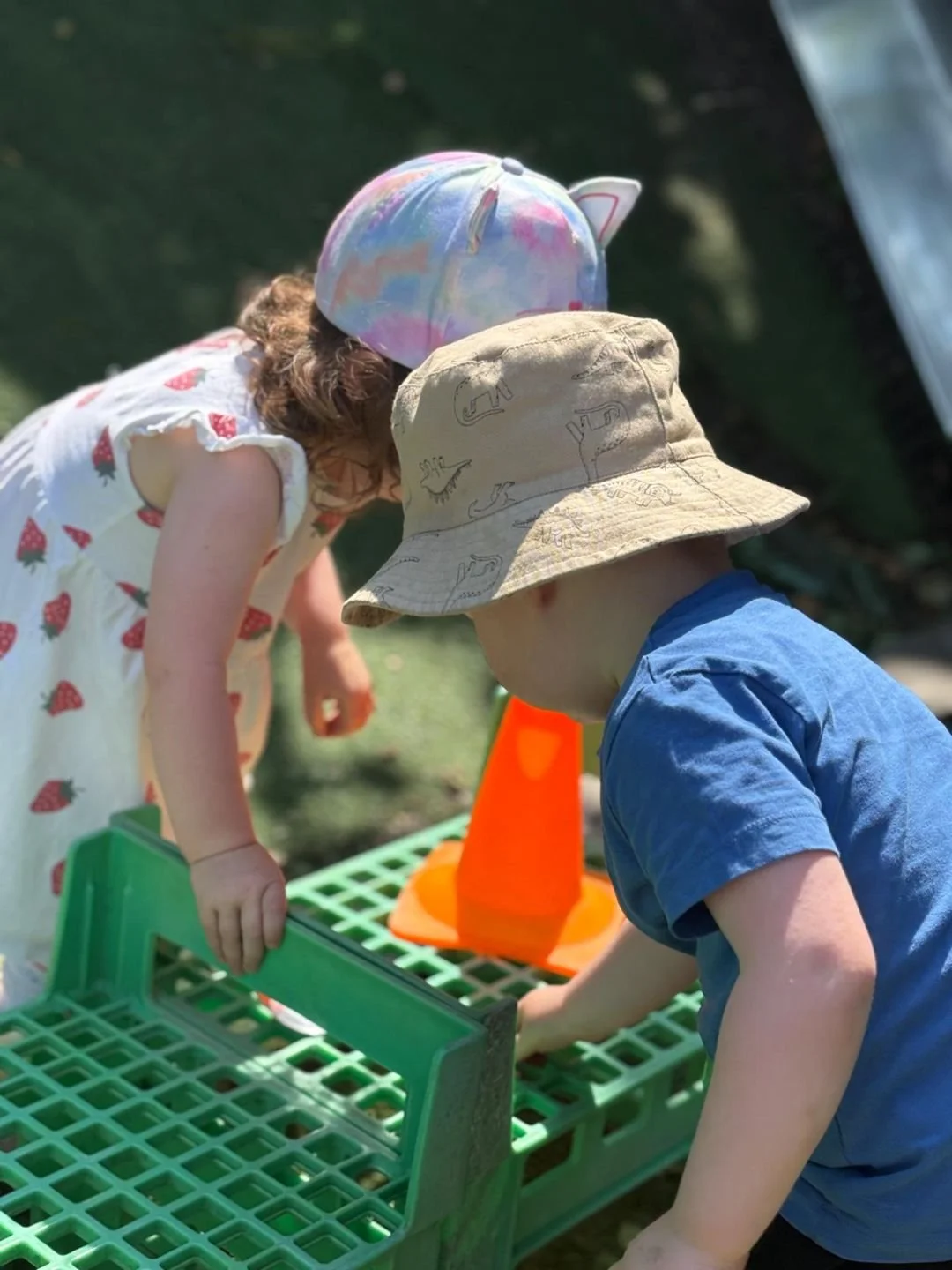 Two young children wearing hats and casual clothes are playing outdoors with a green plastic grid, orange traffic cones, and other toys on the grass.