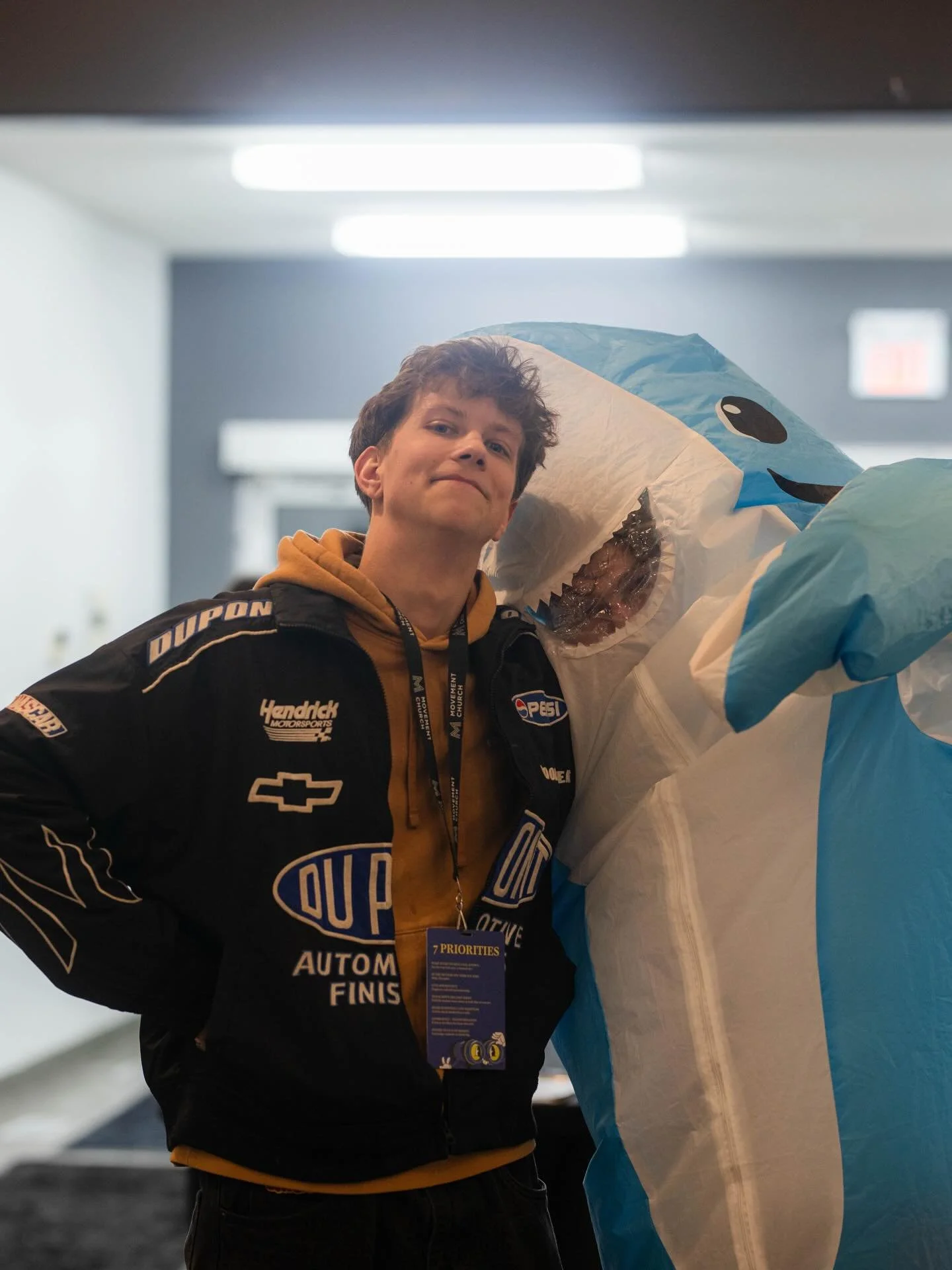 Young man in black racing jacket standing next to a blue and white inflatable shark costume in an indoor setting.