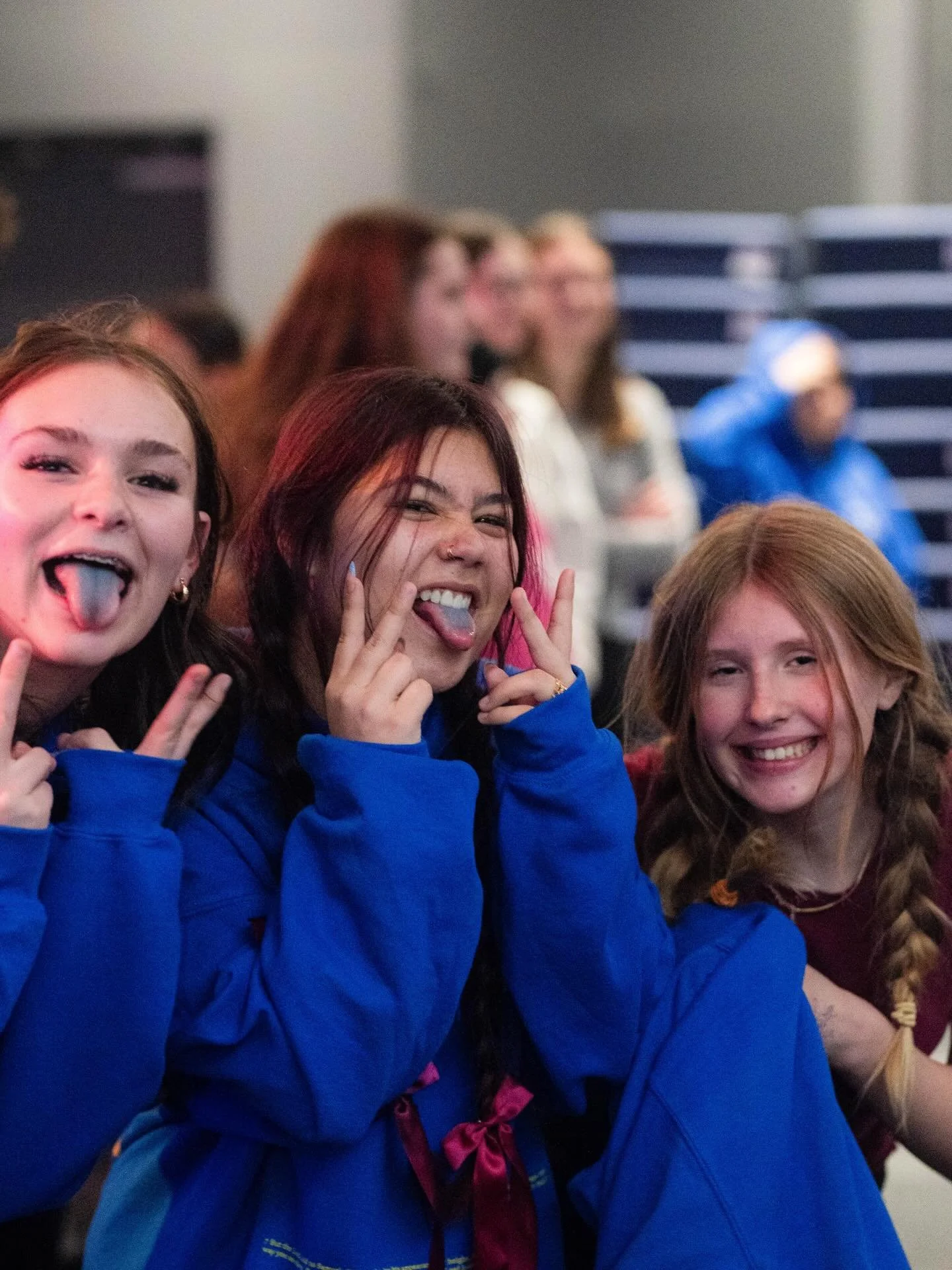 Three young women in blue jackets making silly faces and sticking their tongues out at the camera, indoors with a group of people in the background.