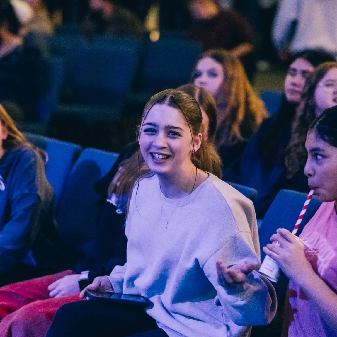 Young woman with brown hair smiling and wearing a gray sweatshirt, sitting in an auditorium surrounded by other people.