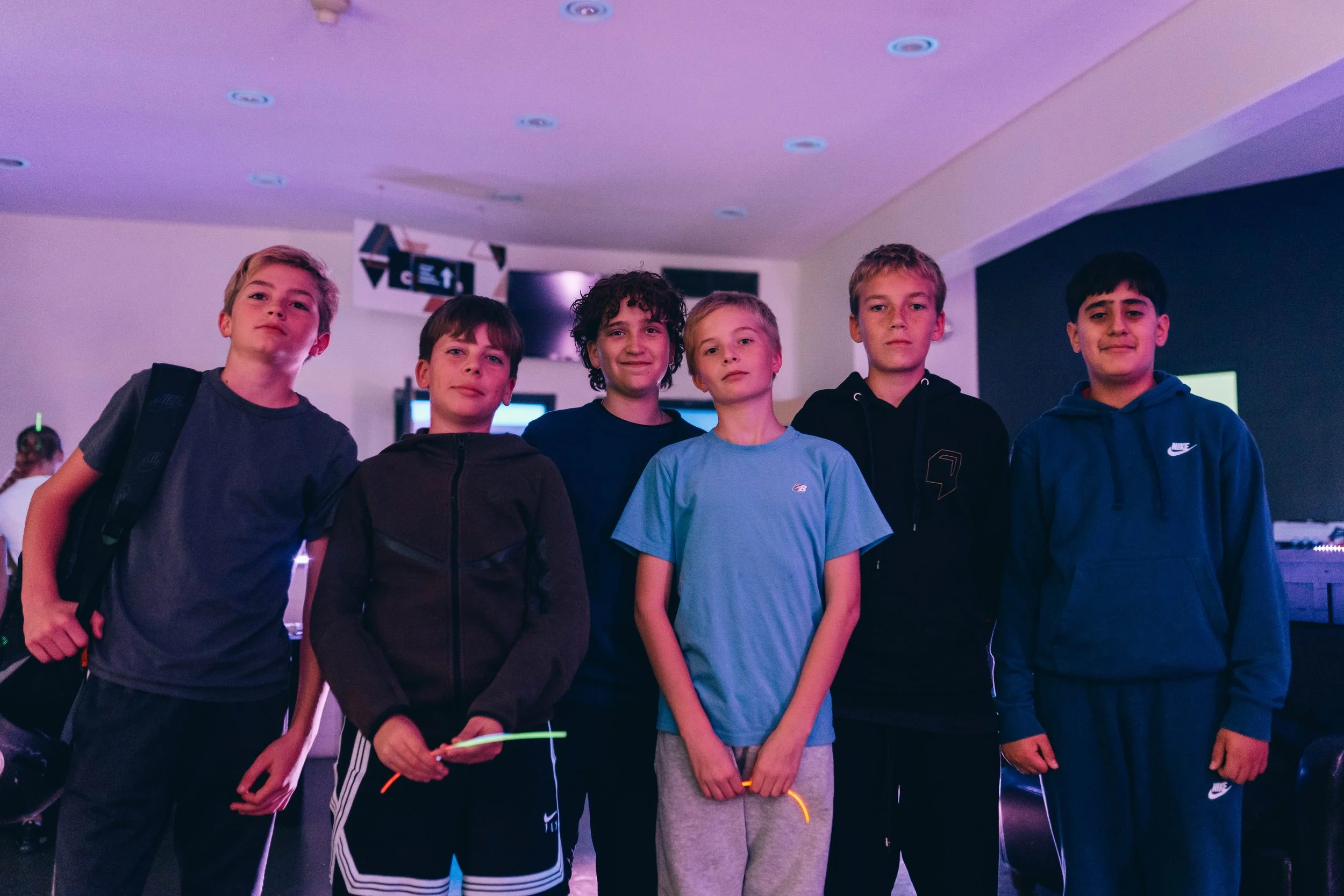 Six boys standing together indoors, some holding glow sticks, in front of a black wall, with a large screen and electronic signs behind them.