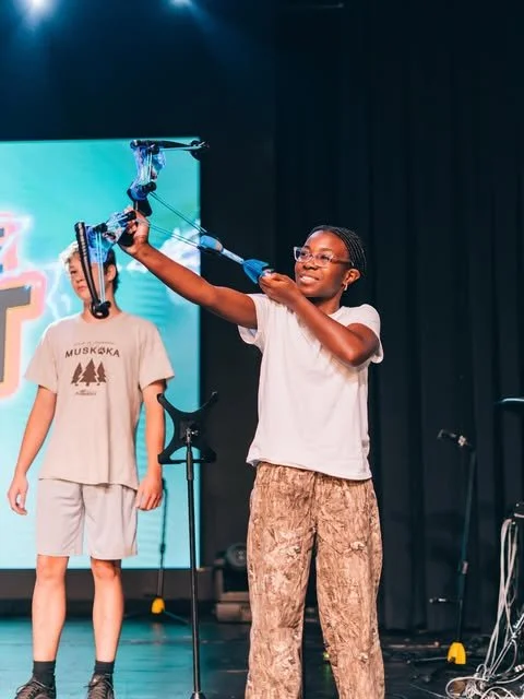 Girl demonstrating a bow and arrow on stage with a boy standing behind her.