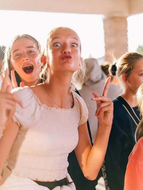 Two young girls at a social gathering, one making a playful pout and pointing, the other smiling broadly with hand raised, with more people in the background.