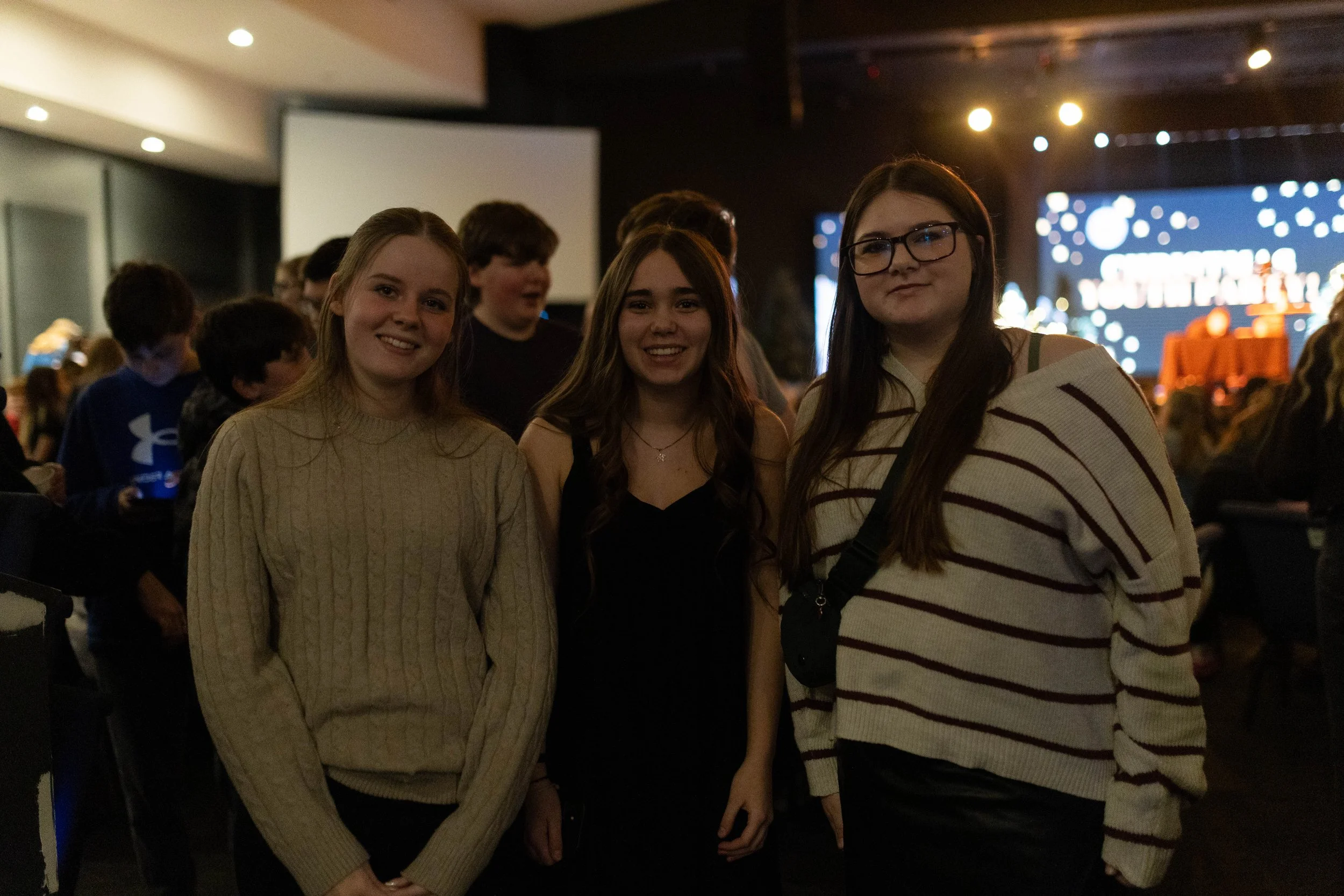 Three young women smiling at the camera at an indoor event with a dark background, stage lights, and a large screen displaying graphics.