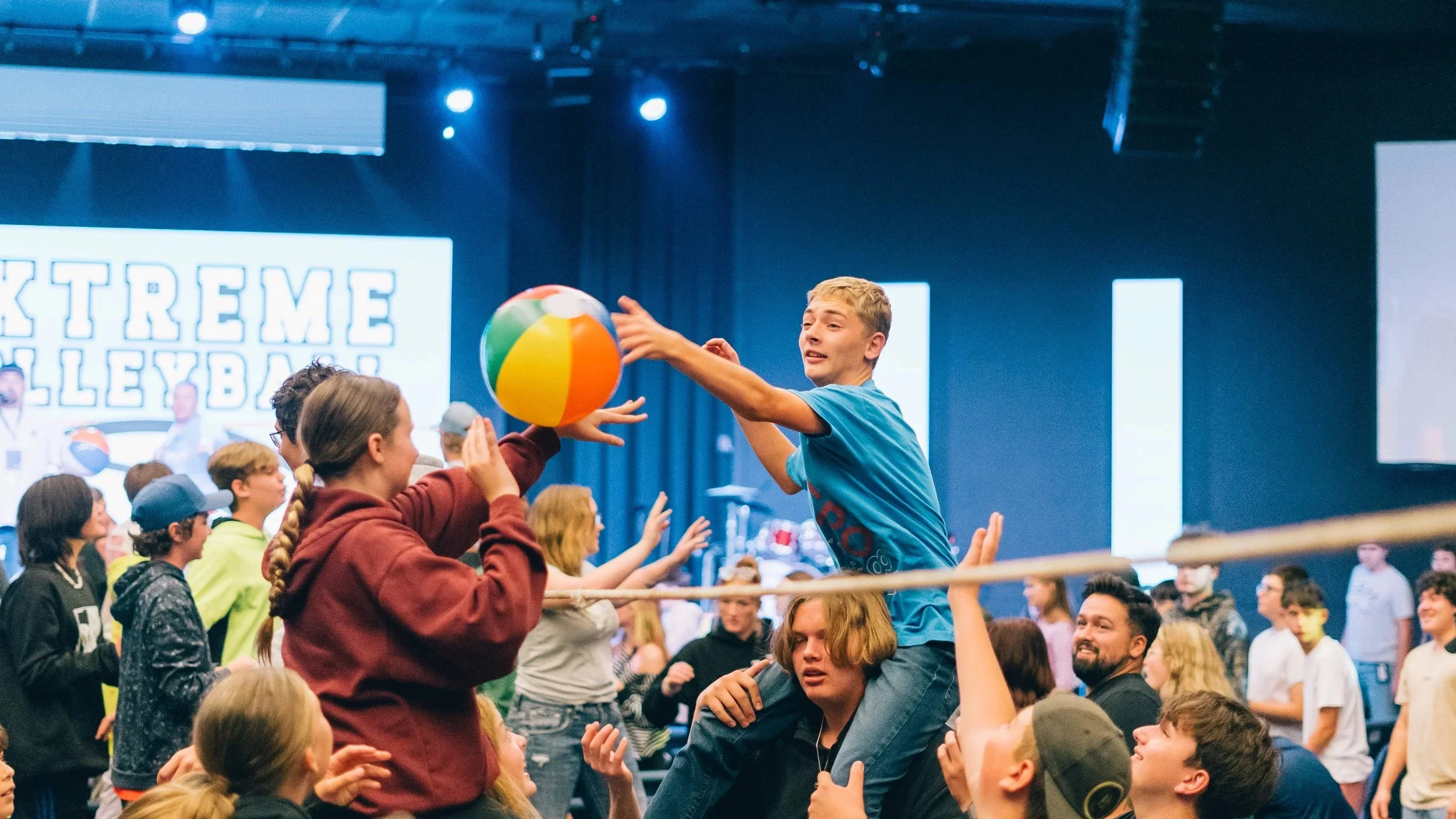 A youth event called 'Extreme Volleyball' with a young man riding on another person's shoulders, reaching to block or catch a colorful beach ball during a lively indoor game, with many other participants and a large digital display behind them.