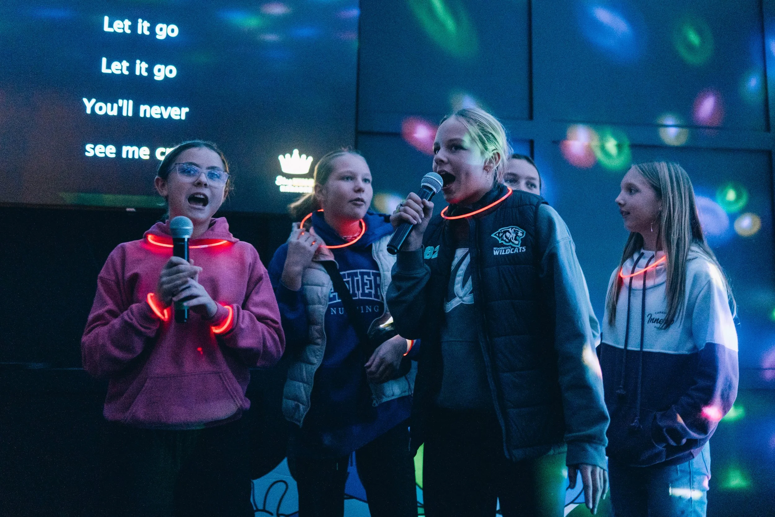 Four young girls singing into microphones on stage, wearing glow necklaces, with lyrics displayed on a large screen behind them.