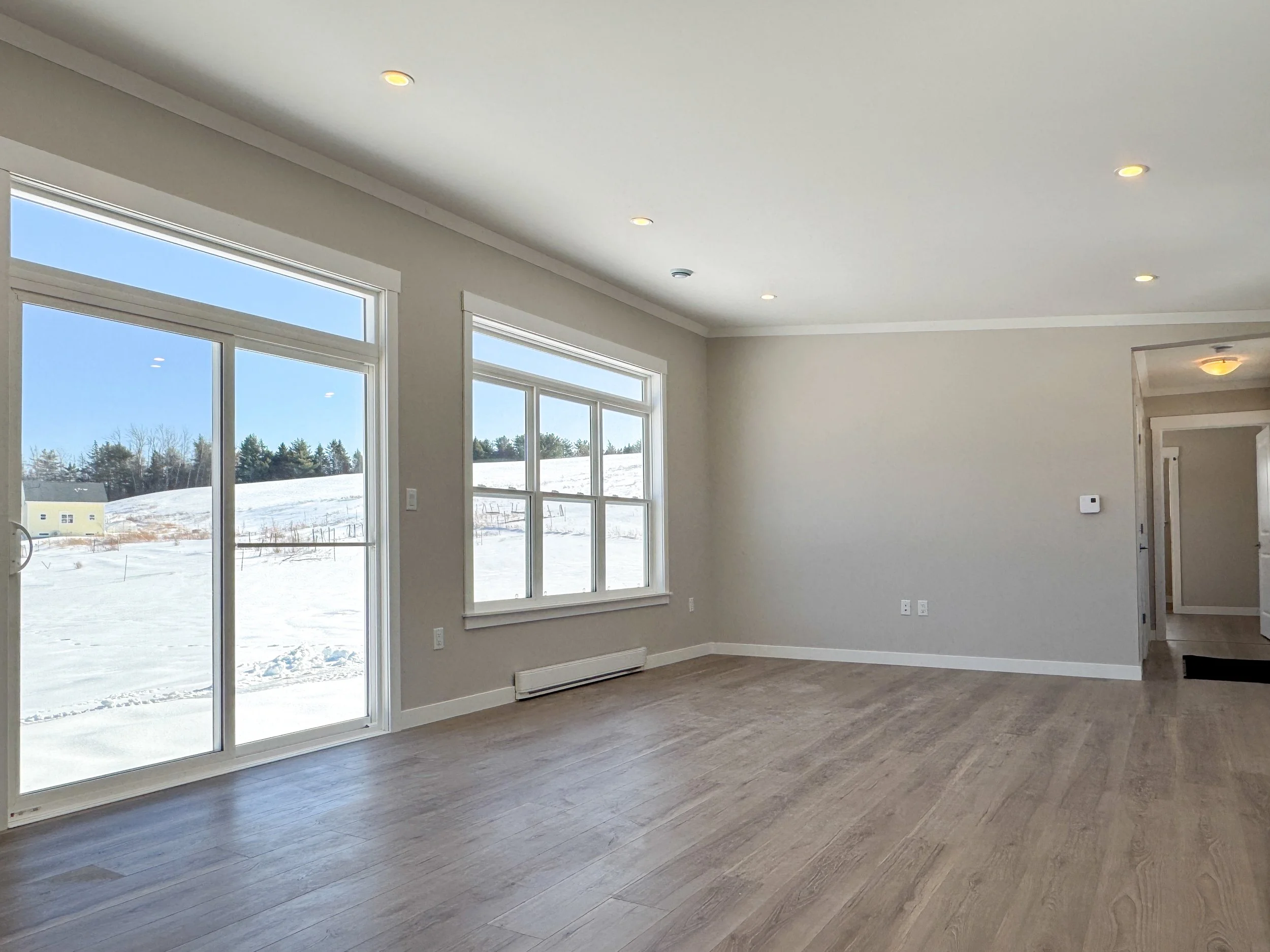 Empty living room with large windows and sliding glass door, showing snowy outdoor landscape, light gray walls, and wooden flooring.