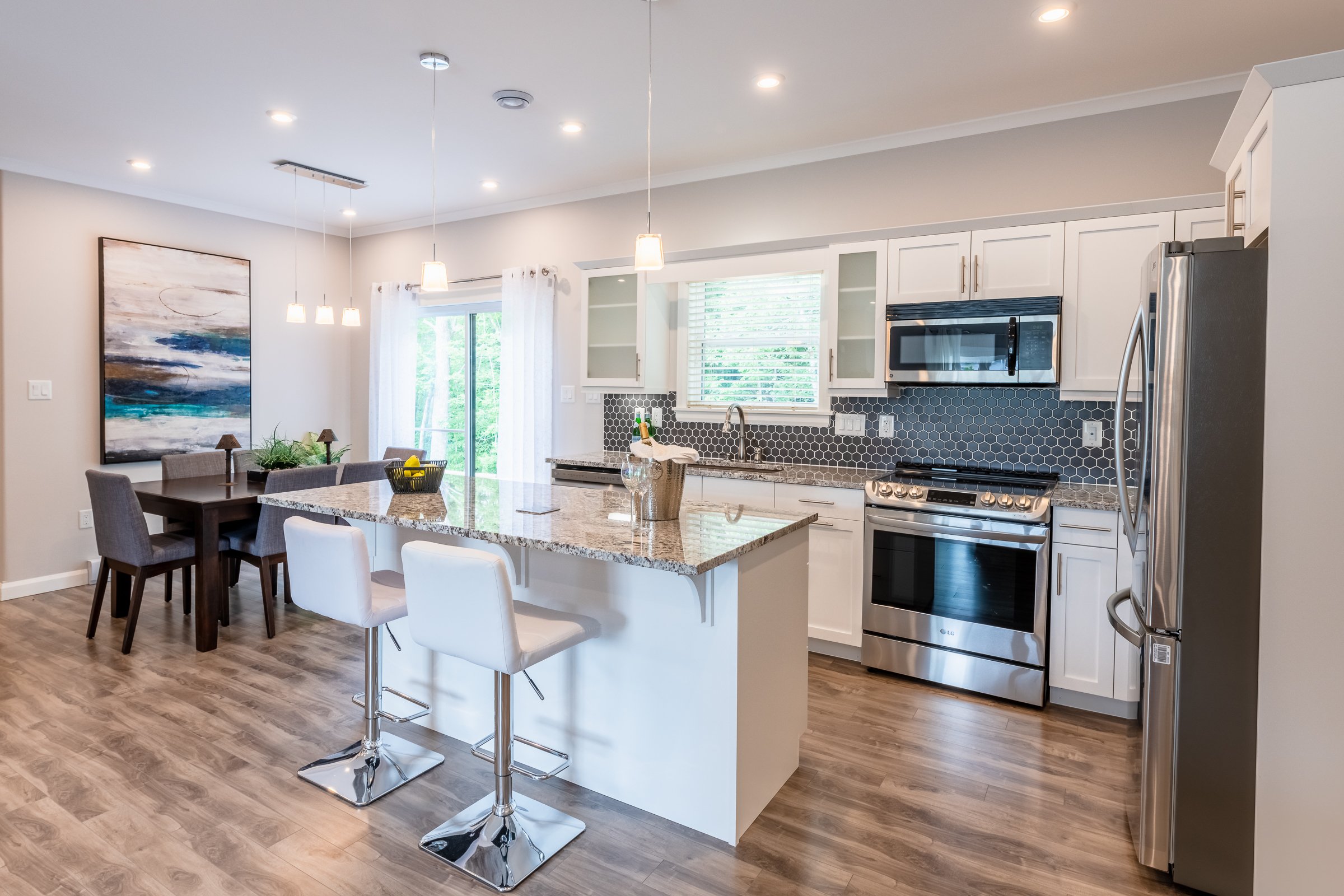 Modern open-concept kitchen with white cabinets, stainless steel appliances, granite island with white bar stools, a dining area with a dark wood table and gray chairs, and wood flooring.