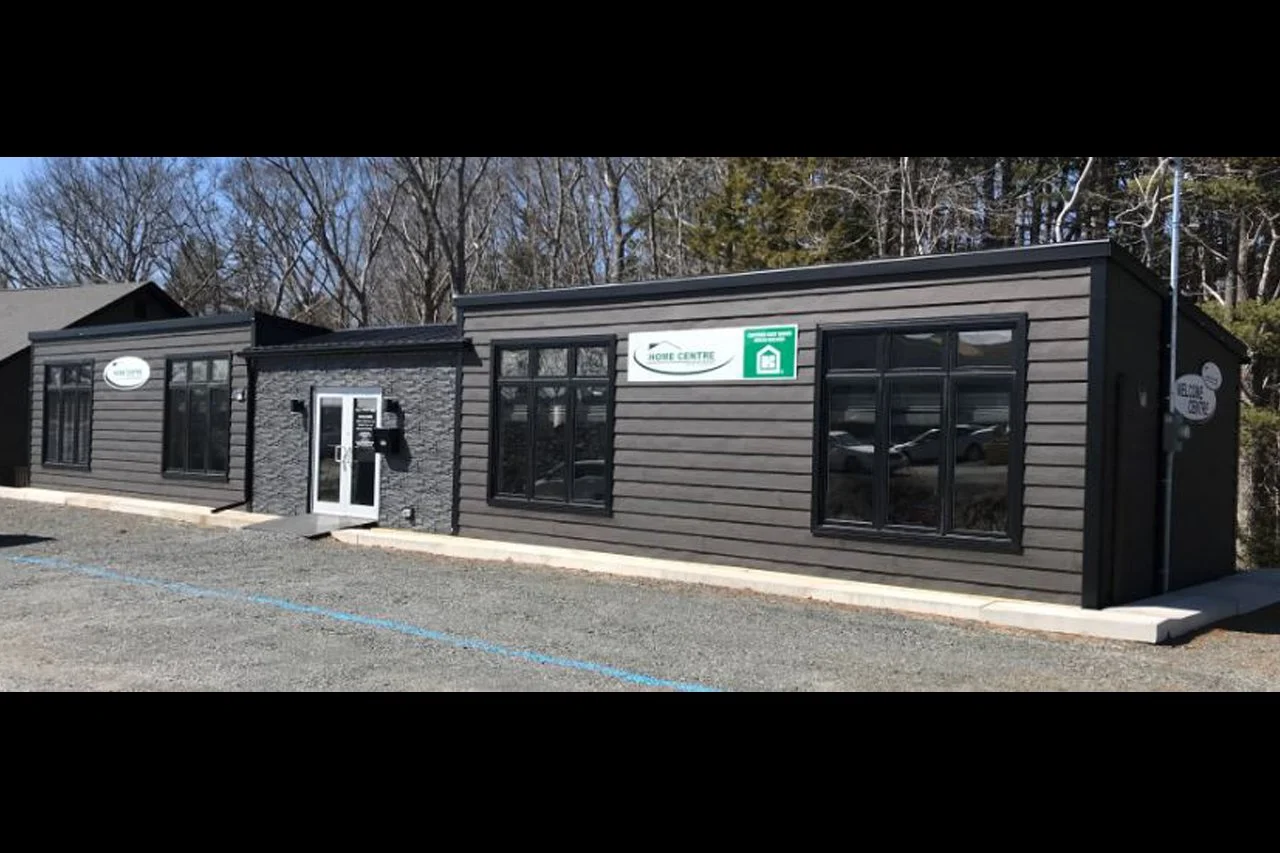 A small, modern commercial building with black siding and multiple large windows, located in a parking lot with a gravel surface and space for parking.