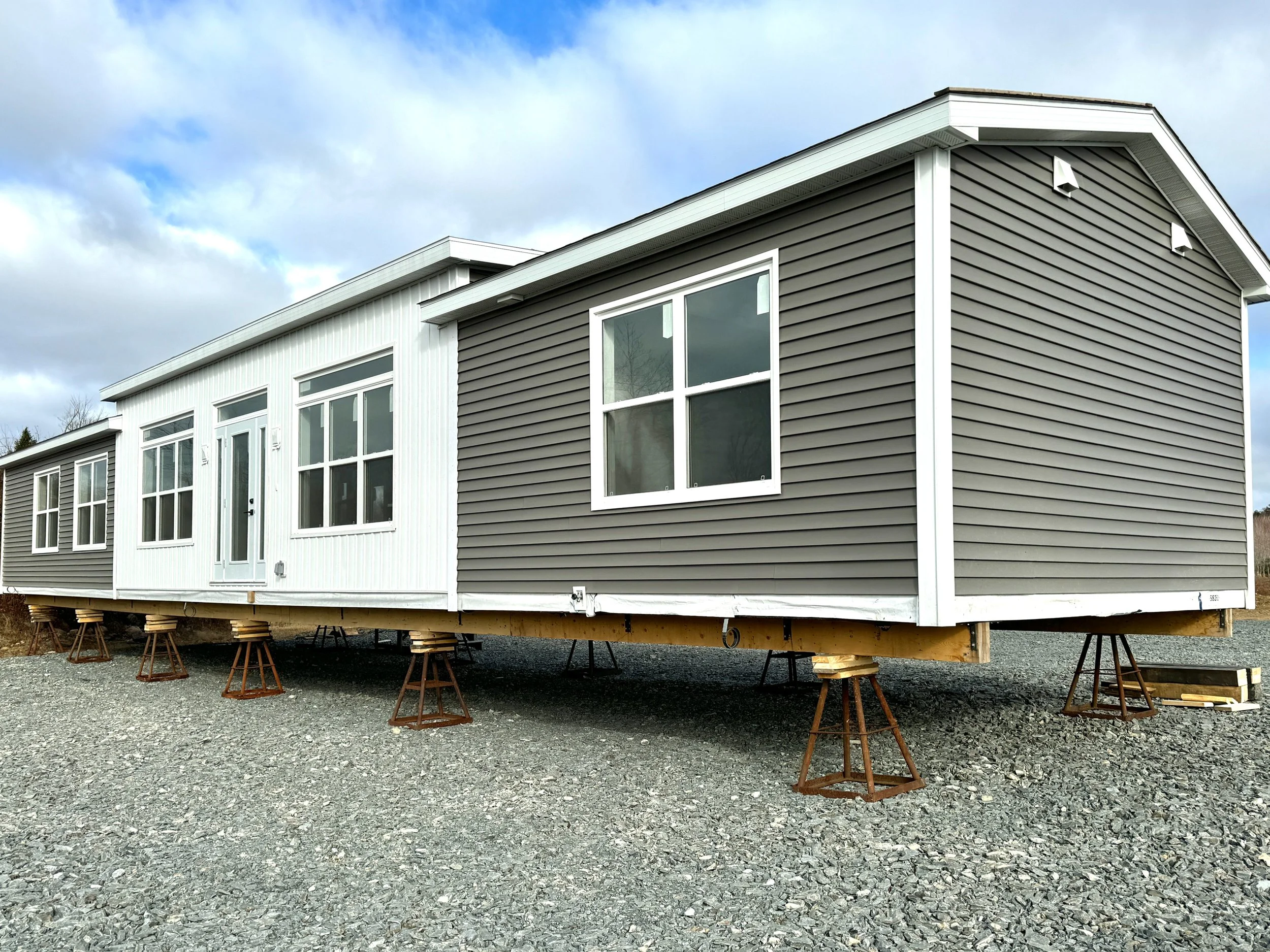 Under construction house with gray and white siding on a gravel lot, supported by jack stands.